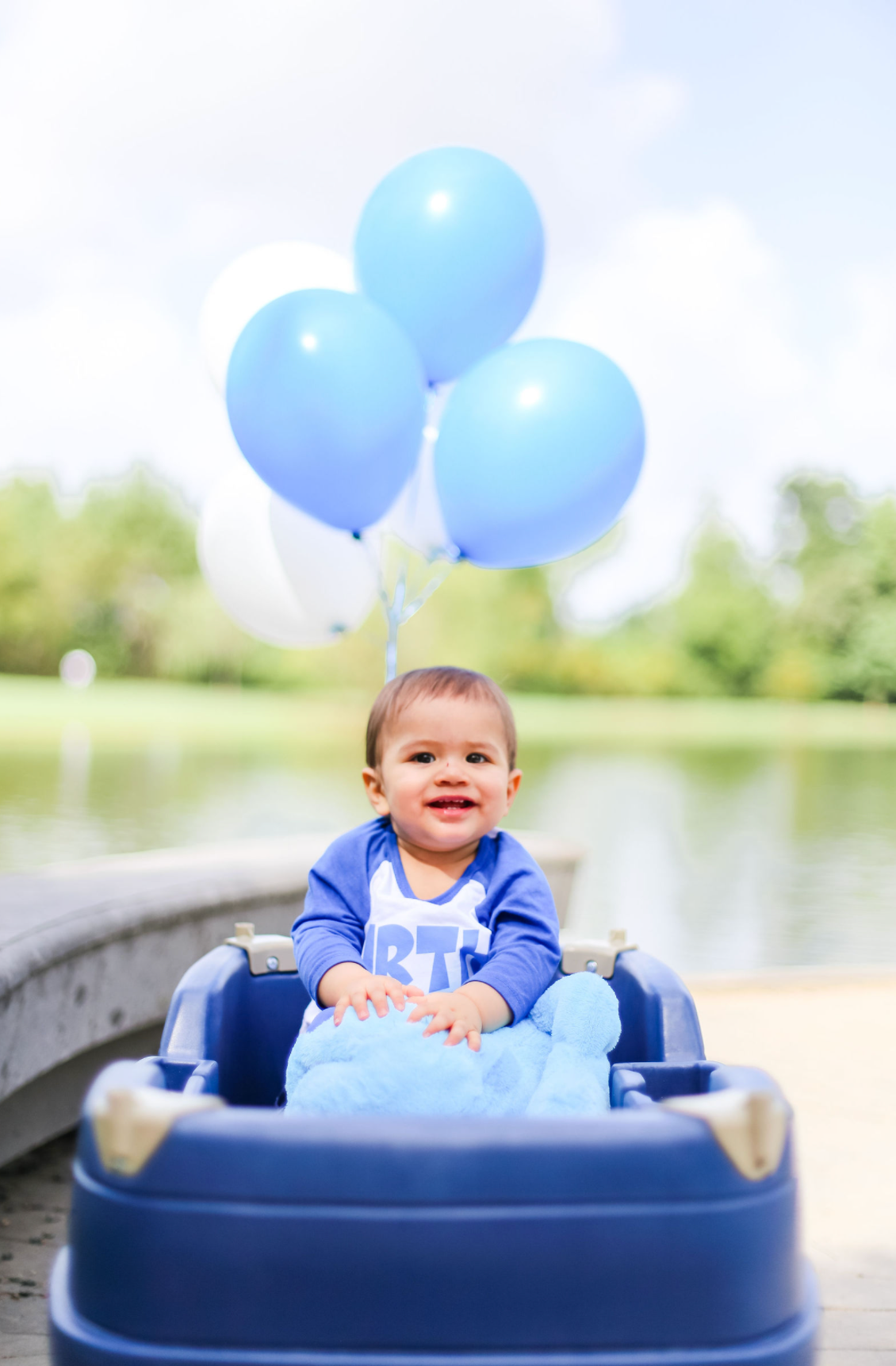 Baby in blue outfit sitting in wagon with balloons near lake.