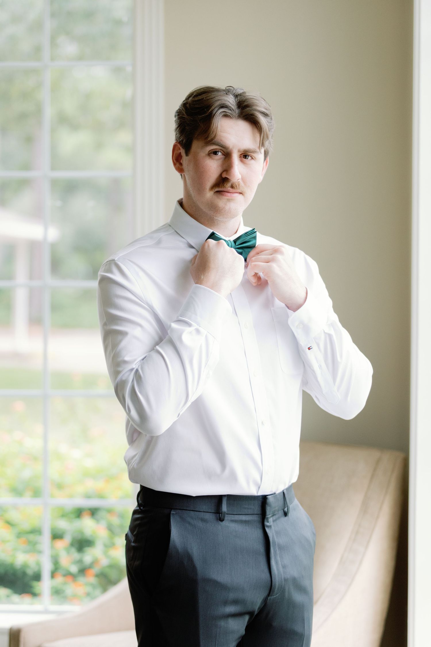 A person in a white dress shirt and dark pants adjusts their bow tie while standing near a bright window.