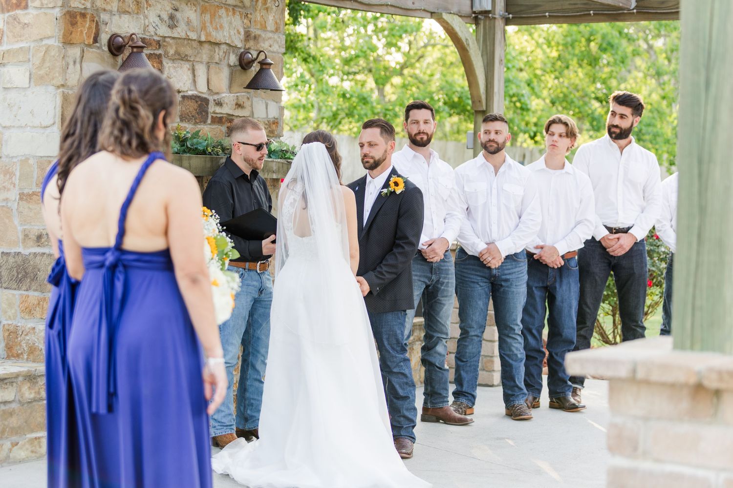 Wedding party gathered under stone pavilion during outdoor ceremony.