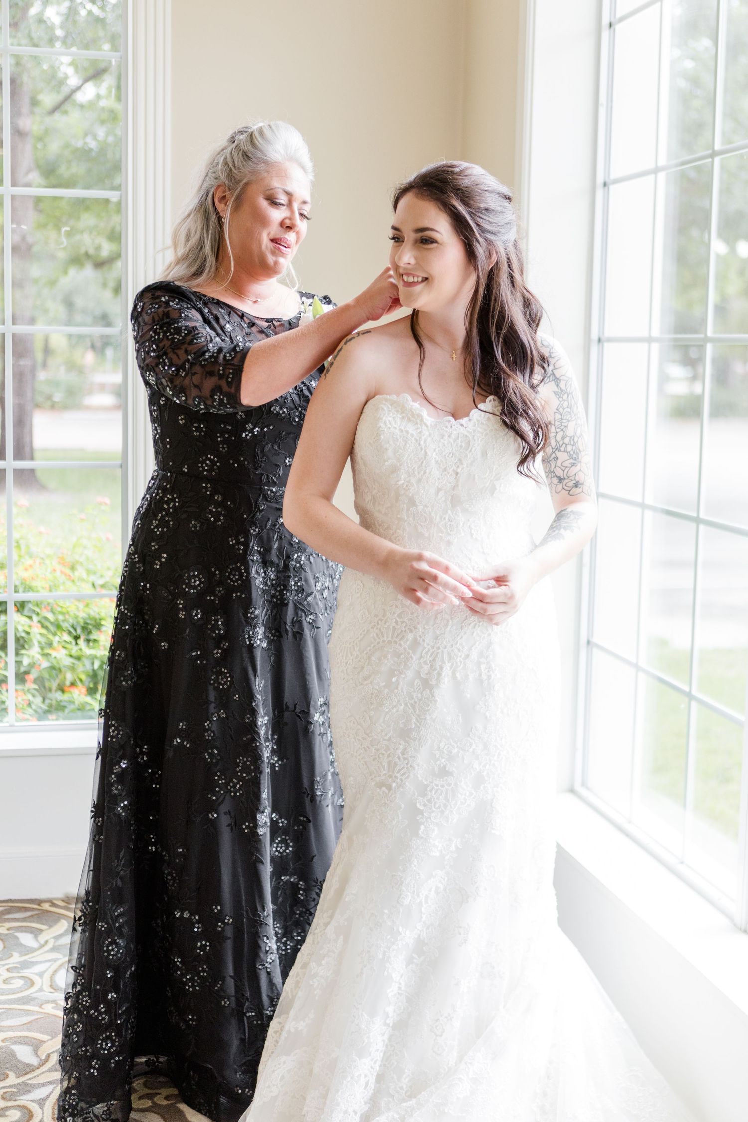 Wedding preparation scene showing bride in white lace dress getting final adjustments near bright windows.