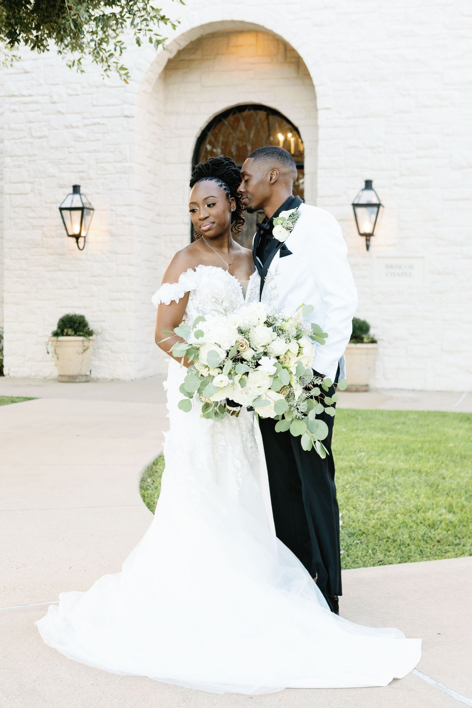 A romantic wedding couple shares intimate moments against a white brick wall with lanterns and lush greenery.