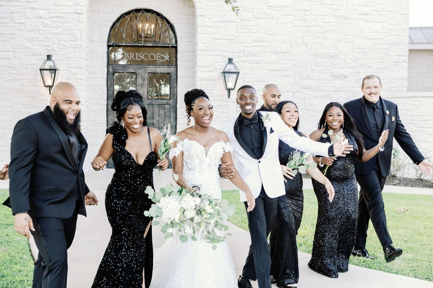 A joyful wedding party in formal attire walking and laughing together outside a white building.