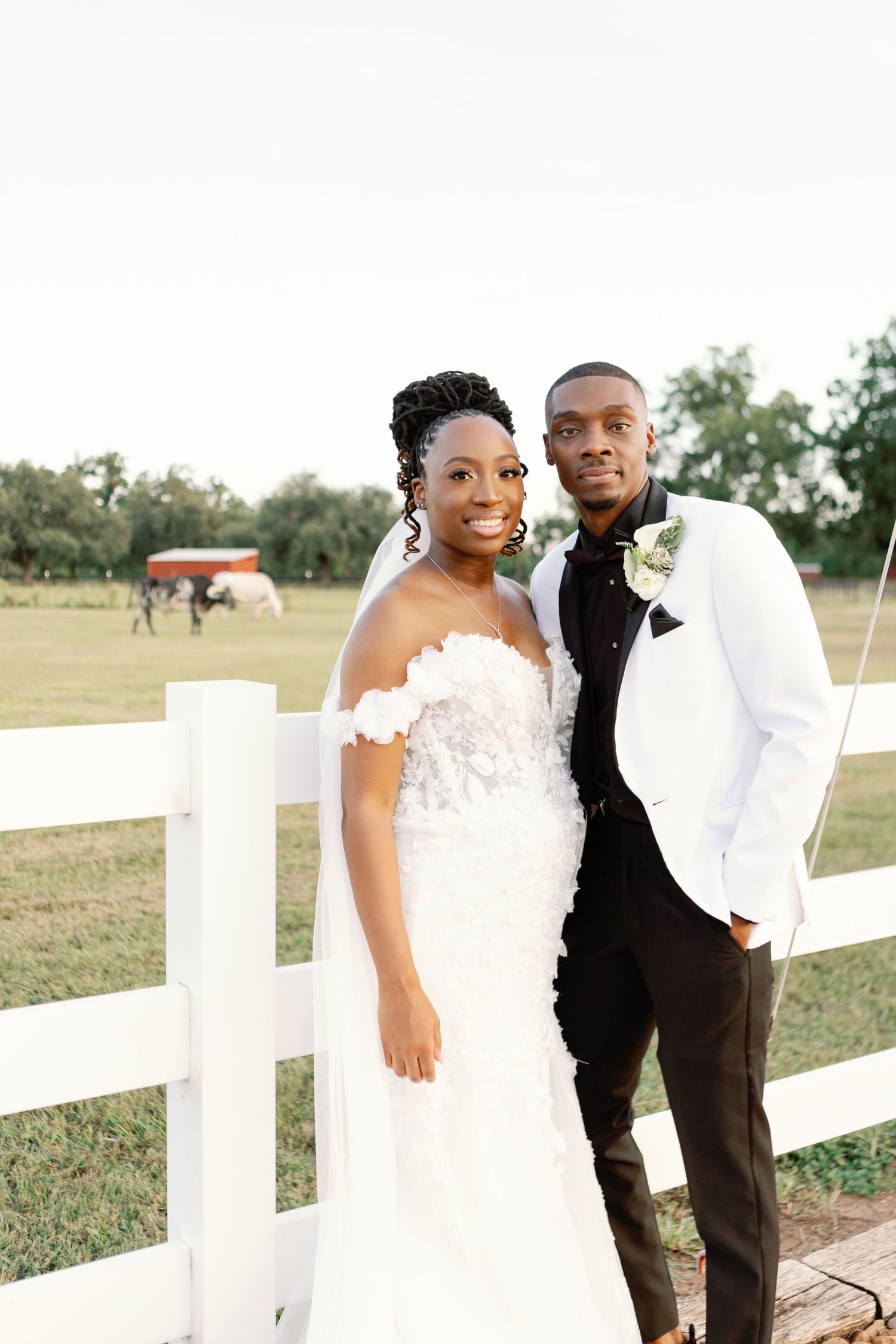 A happy newlywed couple poses together by a white wooden fence at their outdoor wedding ceremony.
