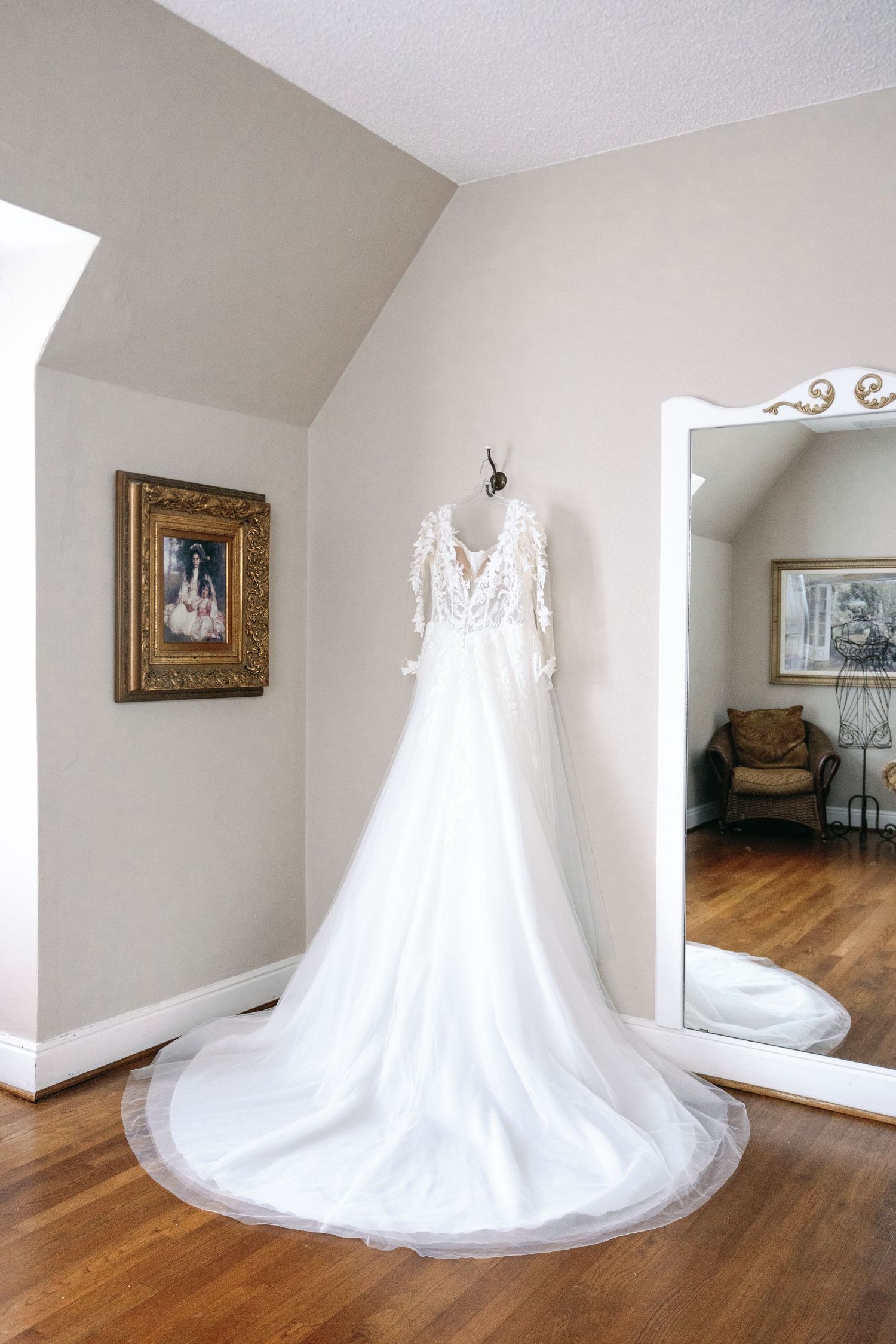 A white lace wedding dress with a long train displayed in a room with hardwood floors and white walls.