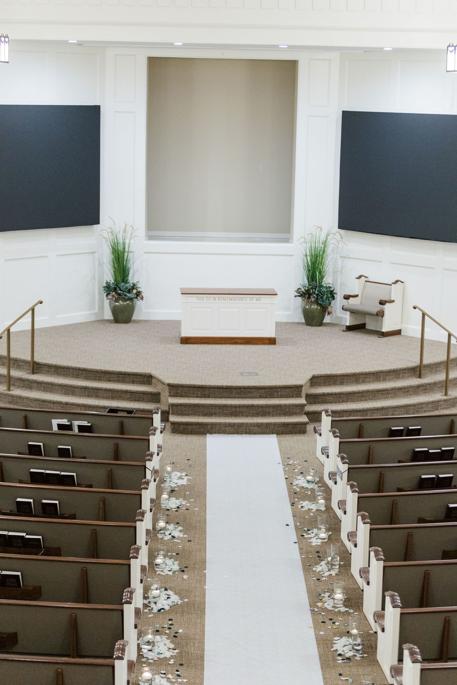 Empty church sanctuary with wooden pews and white walls features decorative floral arrangements along the center aisle.