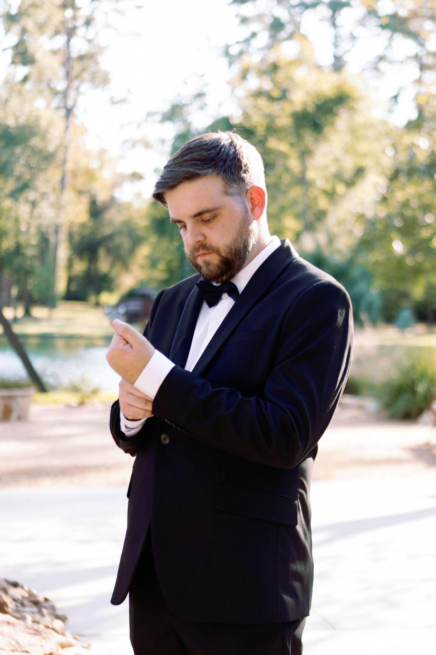 A person in a classic black tuxedo and bow tie adjusts their cuff in an outdoor setting with trees in the background.