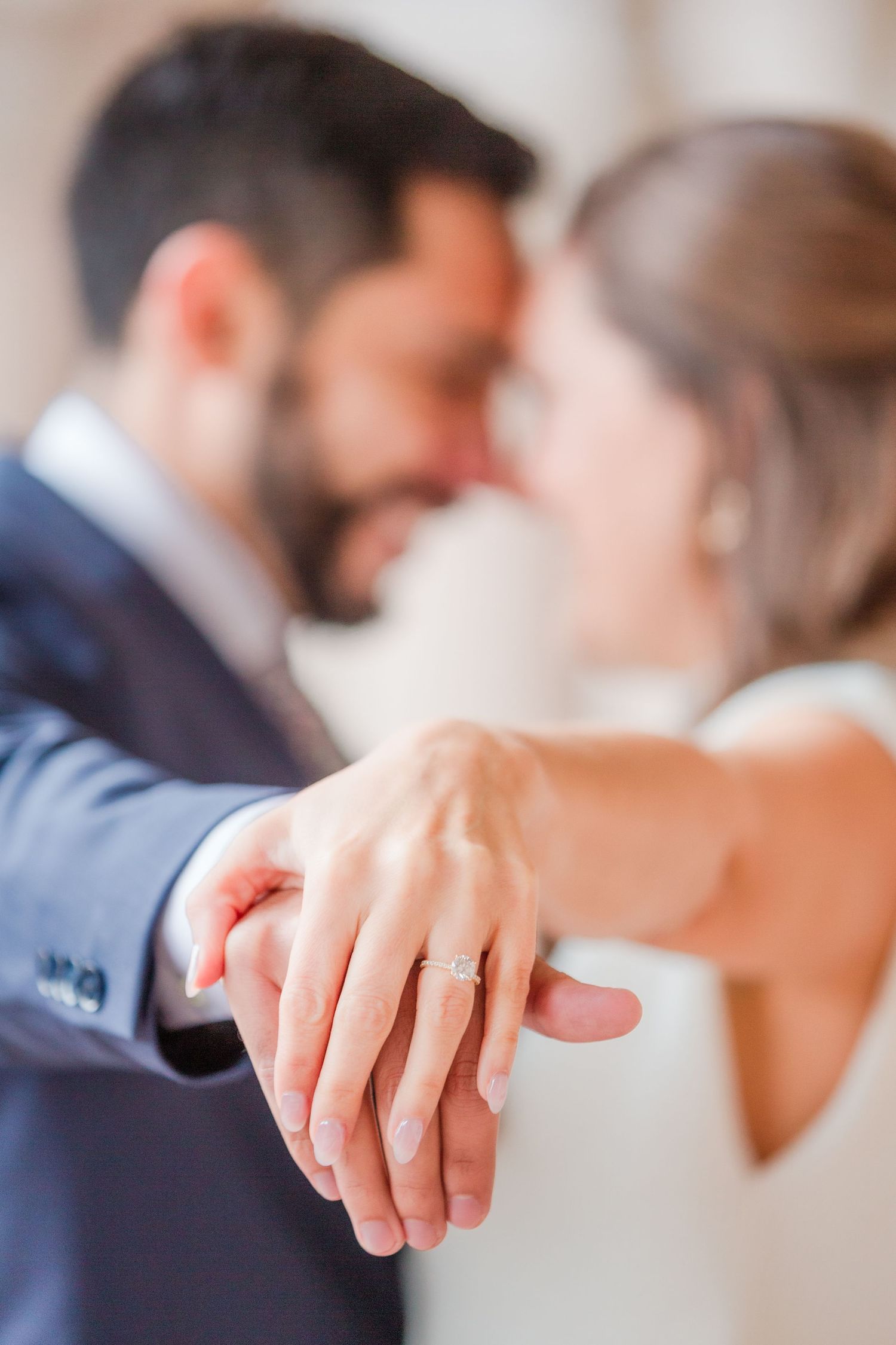 Close up of hands and engagement ring with couple embracing in background.