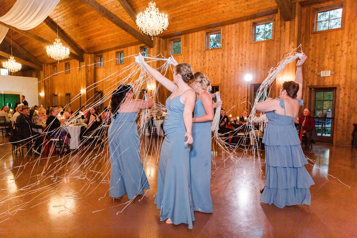 Wedding party celebrates on the dance floor of a rustic wooden venue with elegant chandeliers.