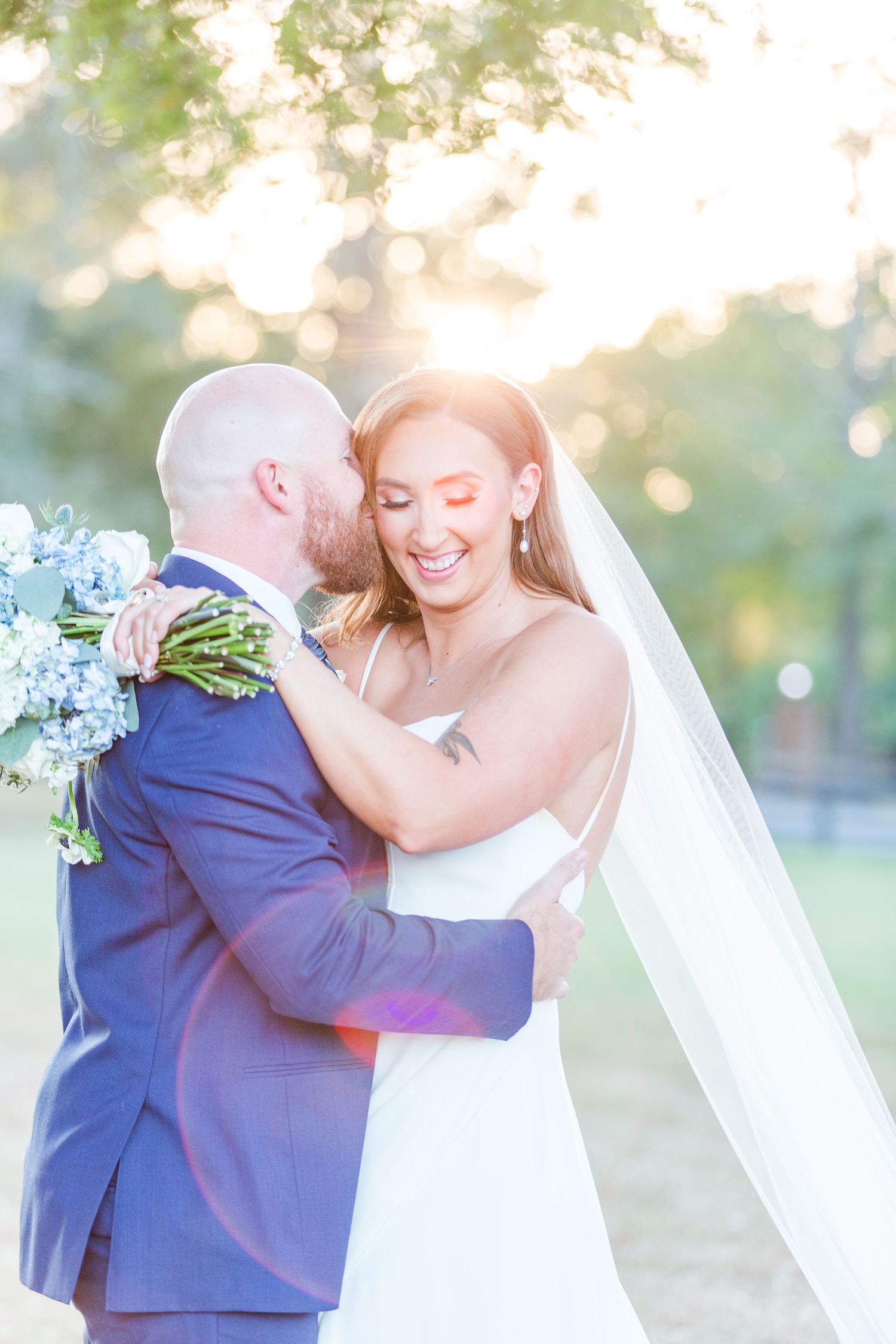 A romantic wedding moment captured in soft sunlight with the couple embracing while wearing blue suit and white dress.