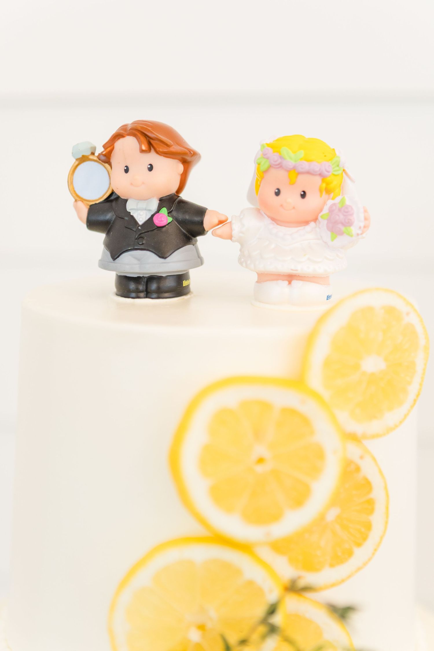Decorative cake topper figurines next to orange slices on a white wedding cake.