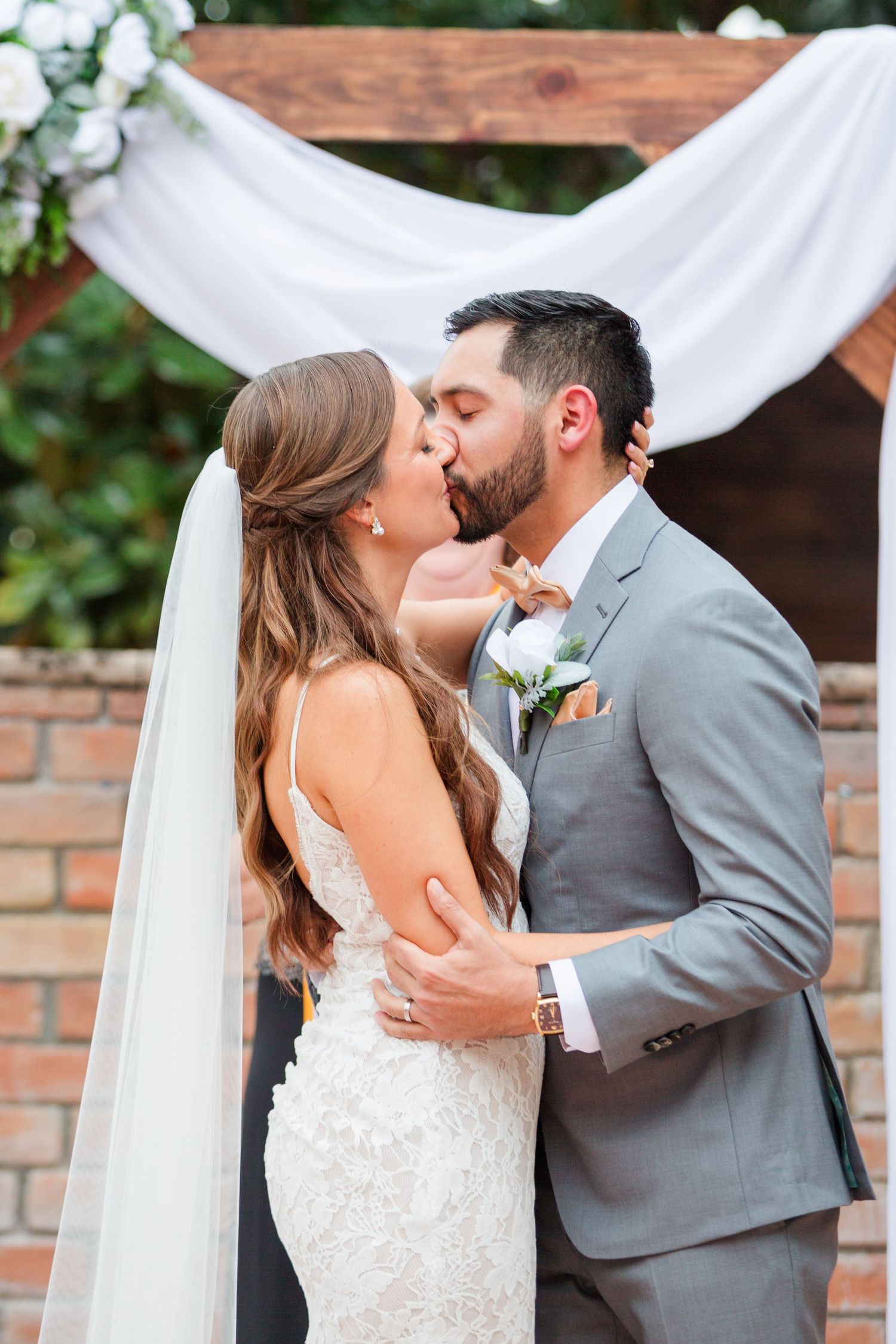 A tender wedding moment captured under an elegant white draped arch with floral decorations.