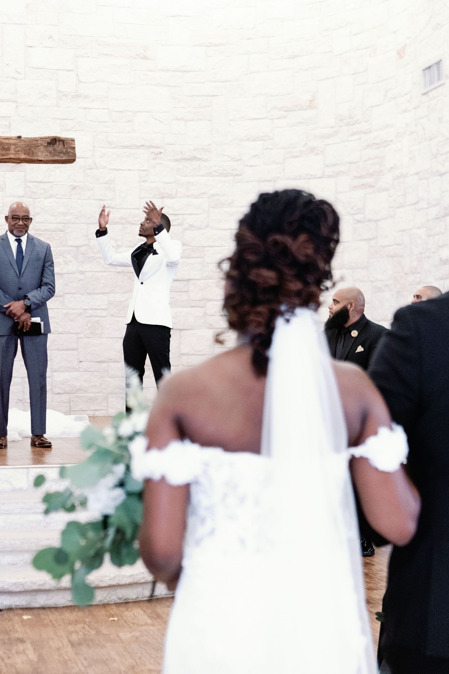 A wedding party gathers against a white stone wall during an outdoor celebration.