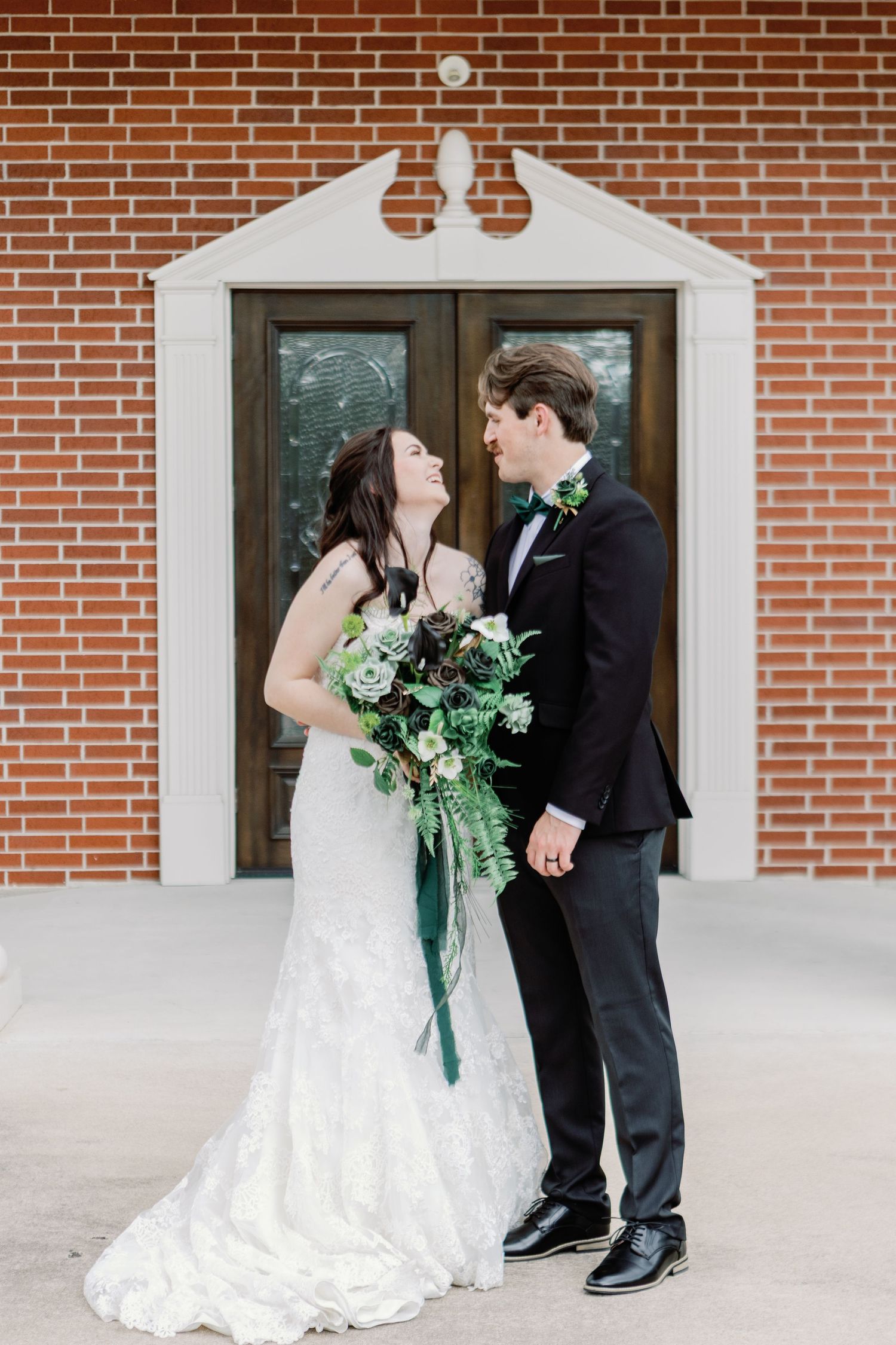 A couple in wedding attire shares a romantic moment against a red brick wall with white columns.