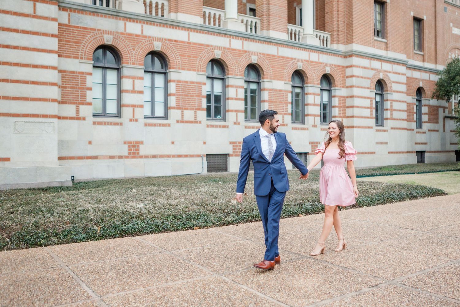 A romantic couple in pink and navy outfits poses for photos along an elegant brick building exterior.