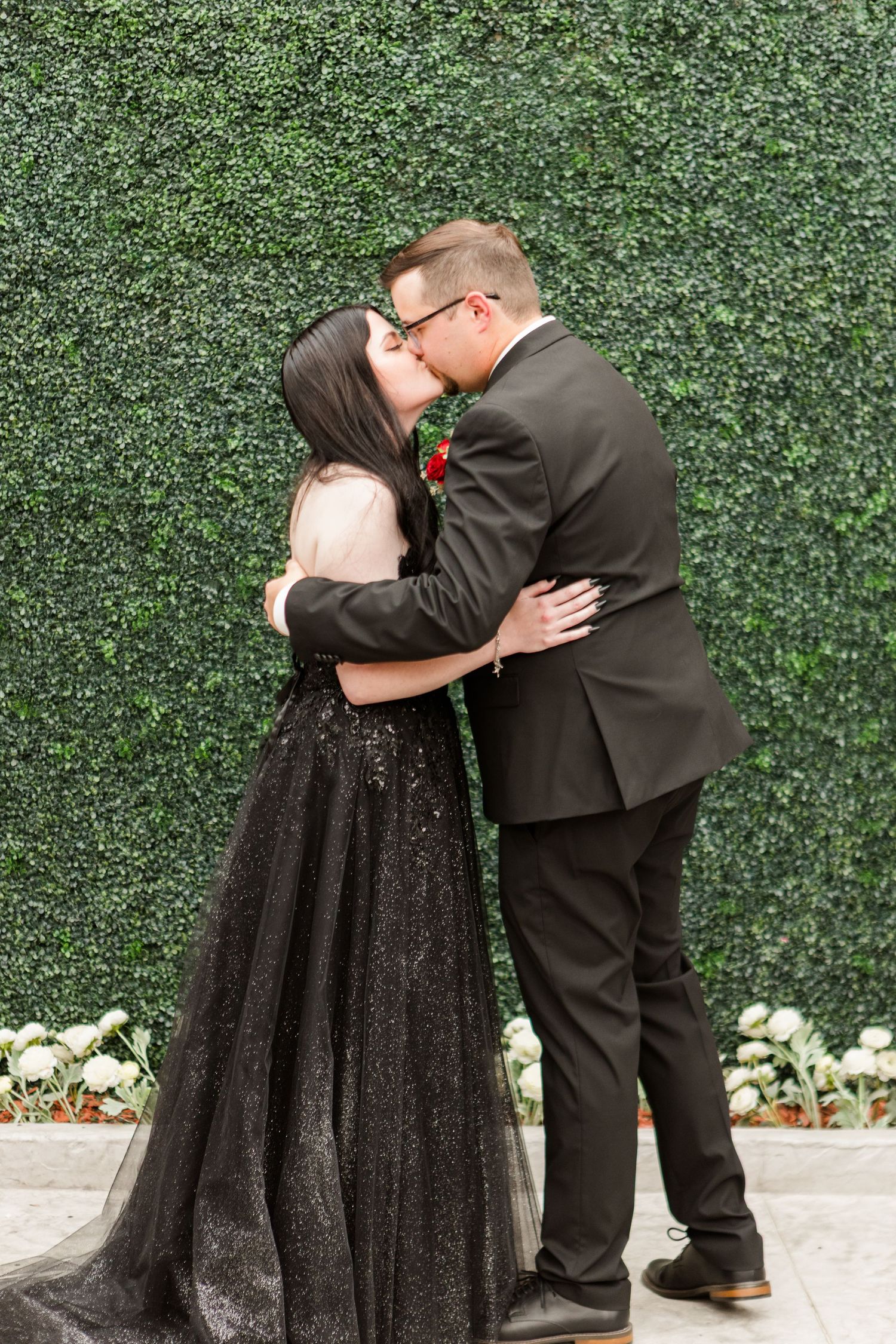 Romantic wedding kiss between couple in black attire against green hedge wall.