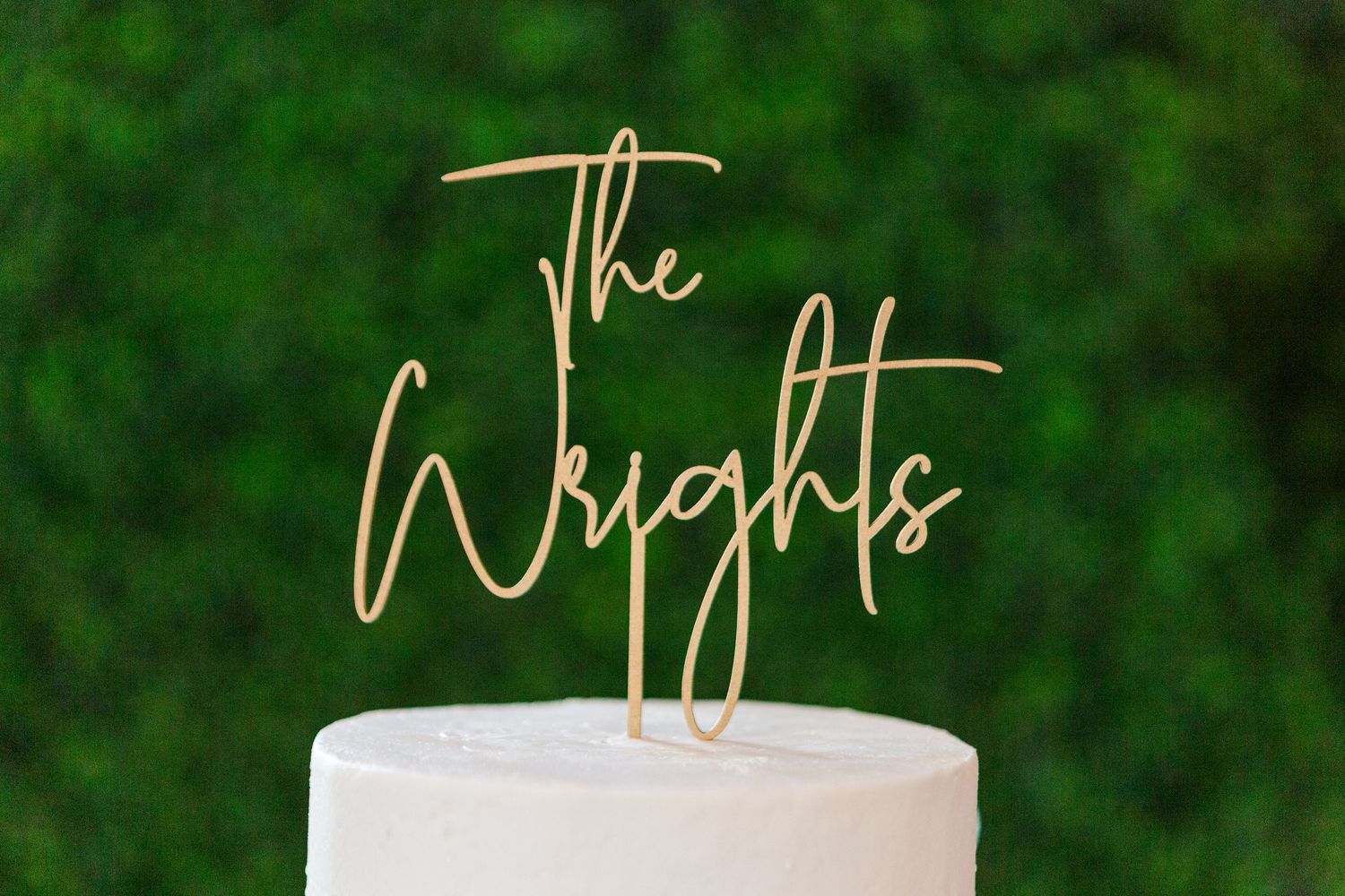 Gold cake topper reading 'The Wrights' on white wedding cake against green backdrop.