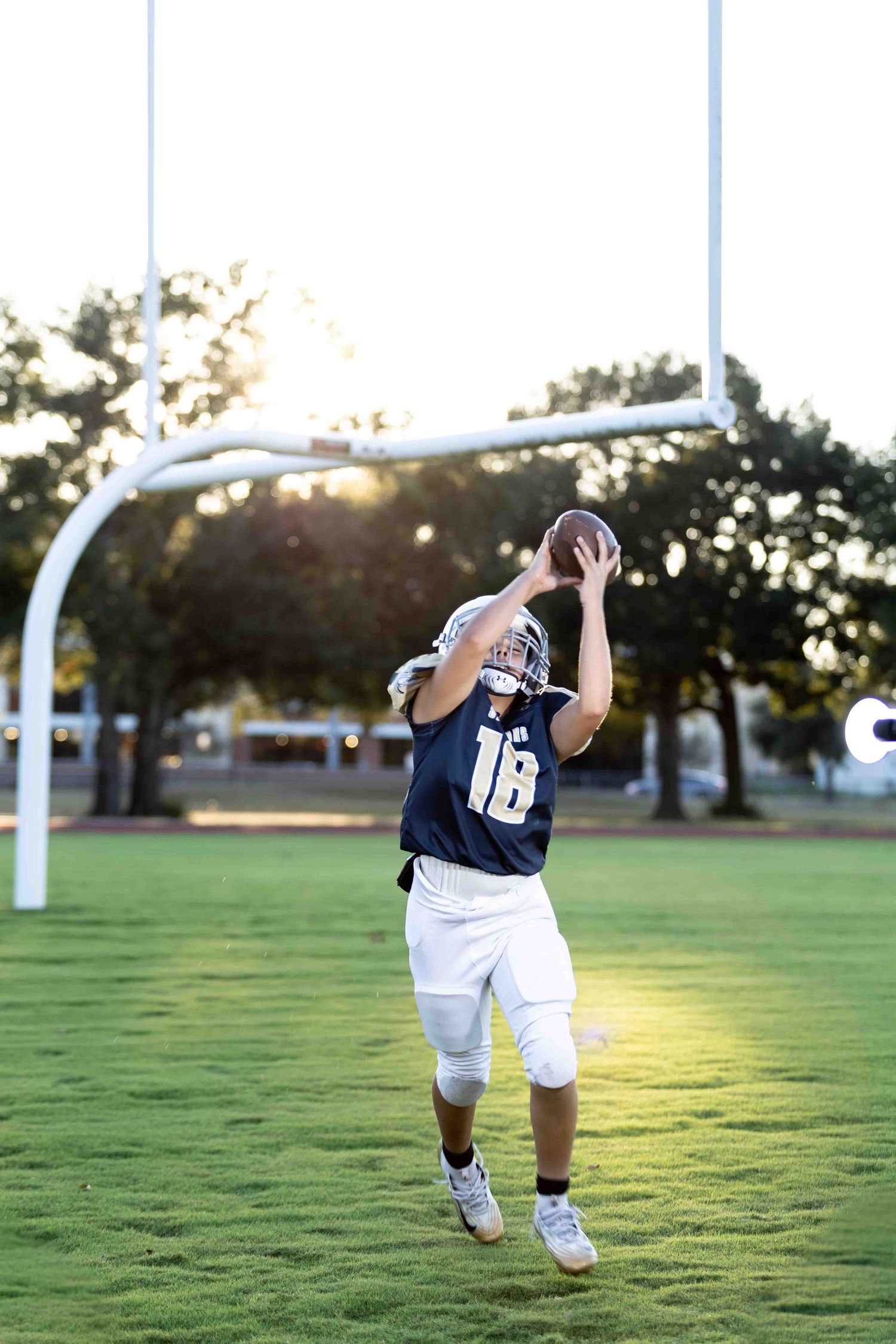 Football player in navy jersey number 18 practices diving catch on grassy field during sunset.