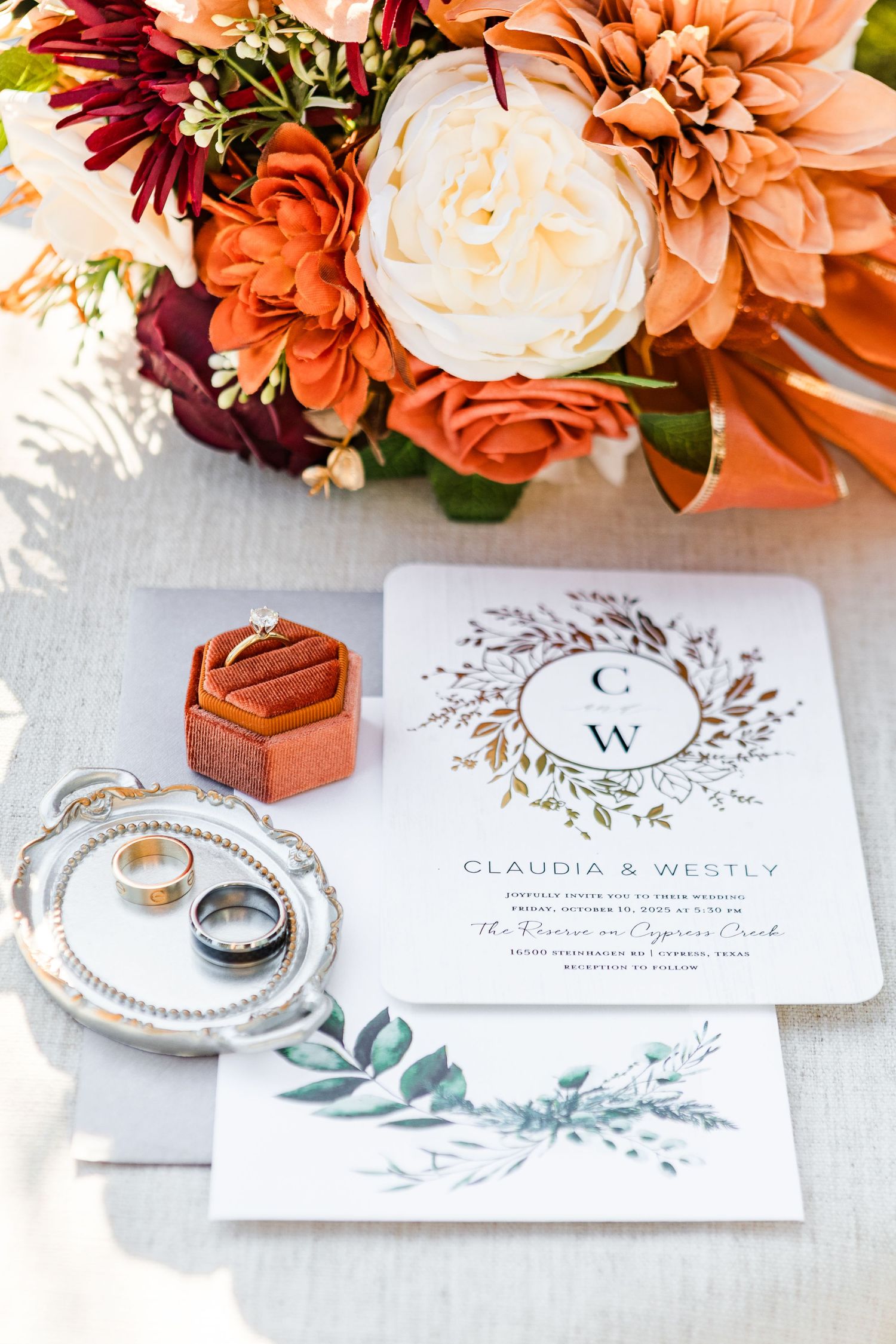 Wedding details featuring autumn-colored floral arrangement with invitation and rings on silver tray.