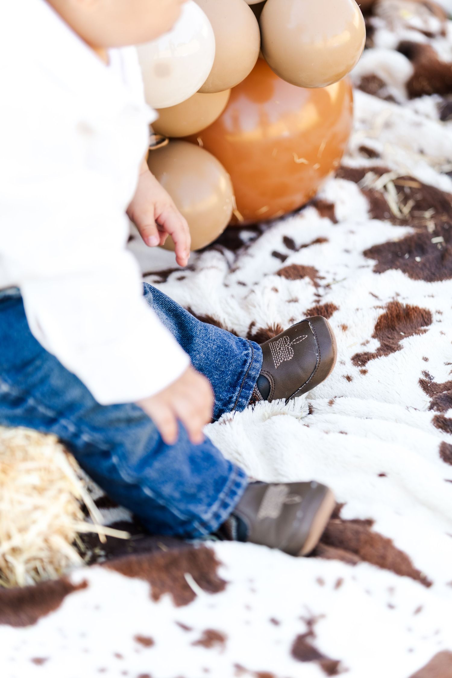 Close-up view of small shoes and jeans on white fabric near hay and balloons.
