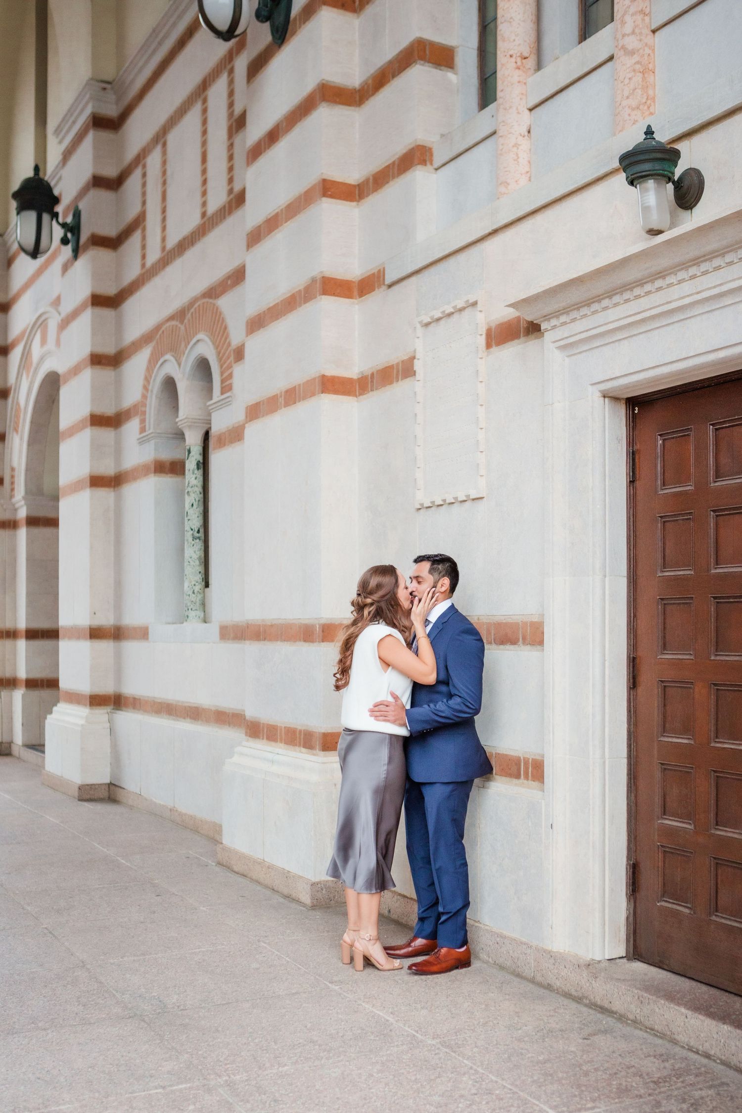 Couple in formal attire embracing against a white wall with ornate architectural details.