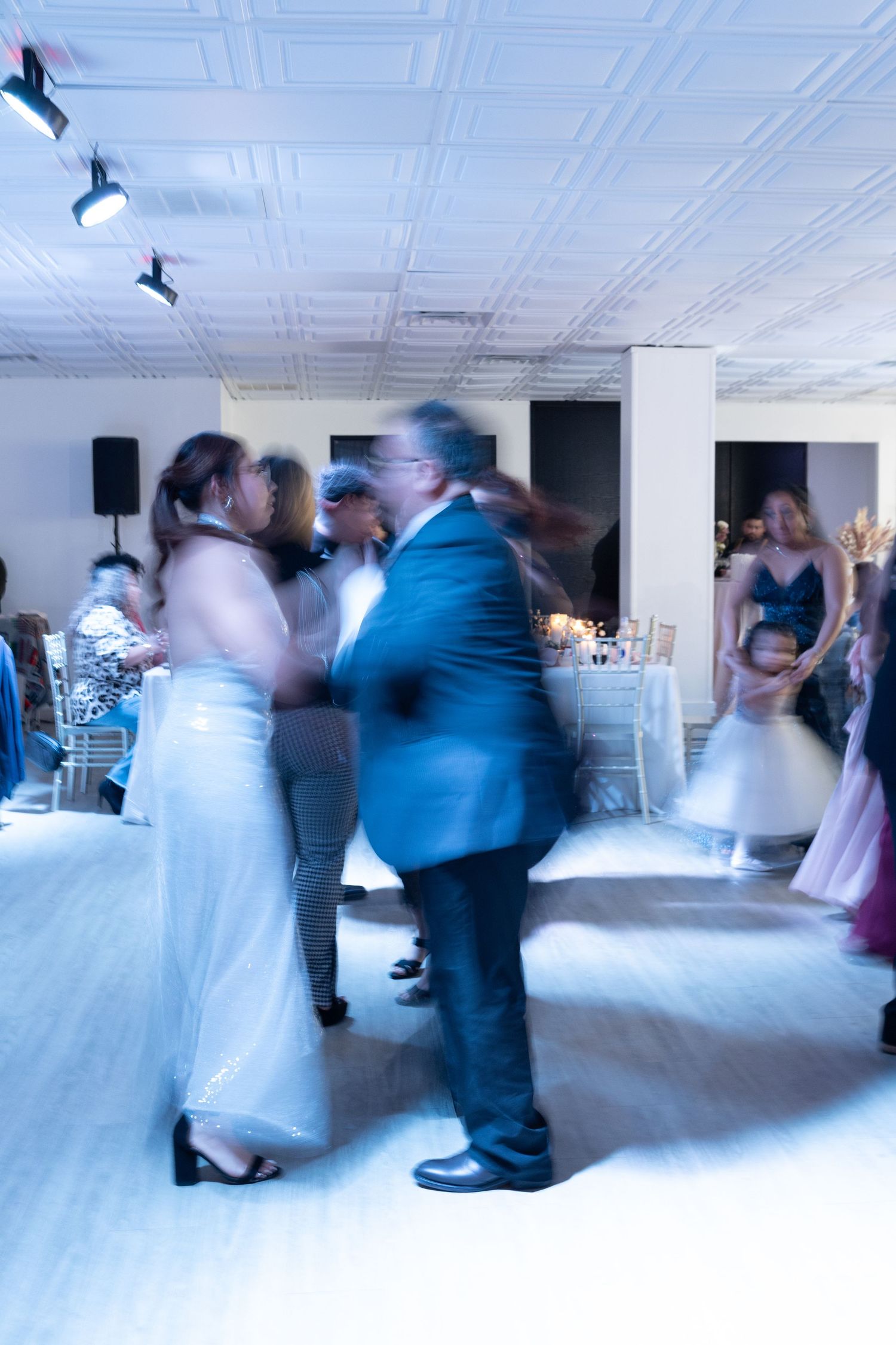 Motion-blurred dancers twirling on a white dance floor under ornate ceiling lighting.