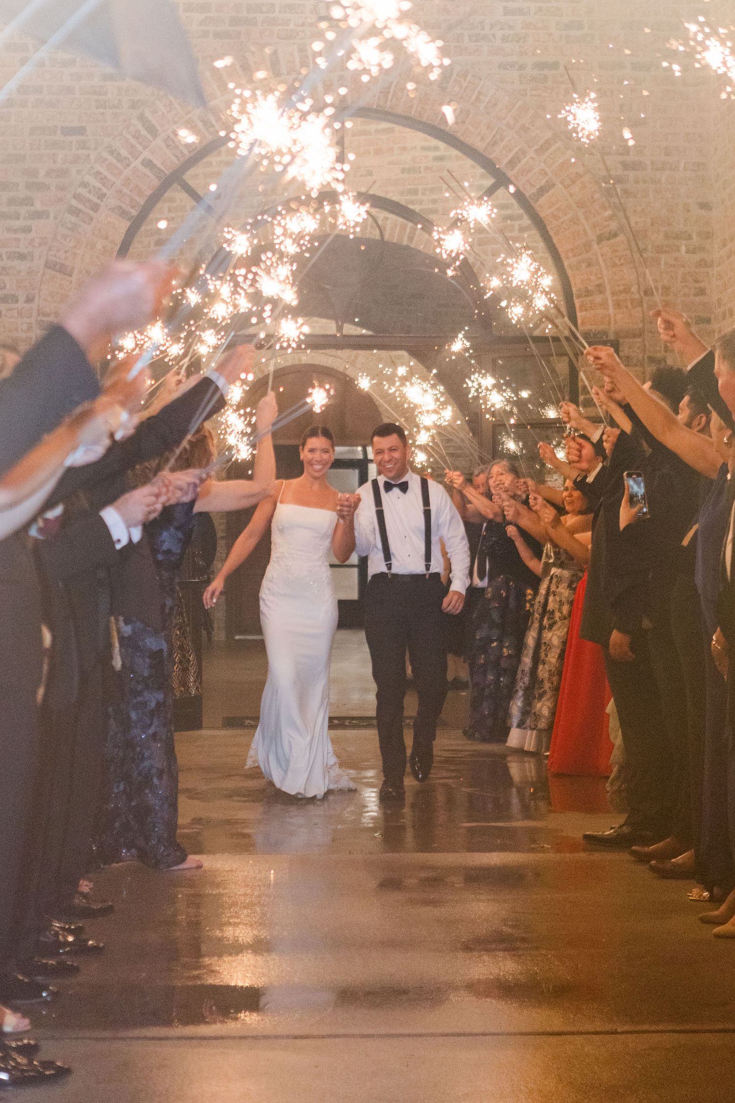 Newlyweds exit their wedding reception through a sparkler tunnel lined with guests in a brick-walled venue.