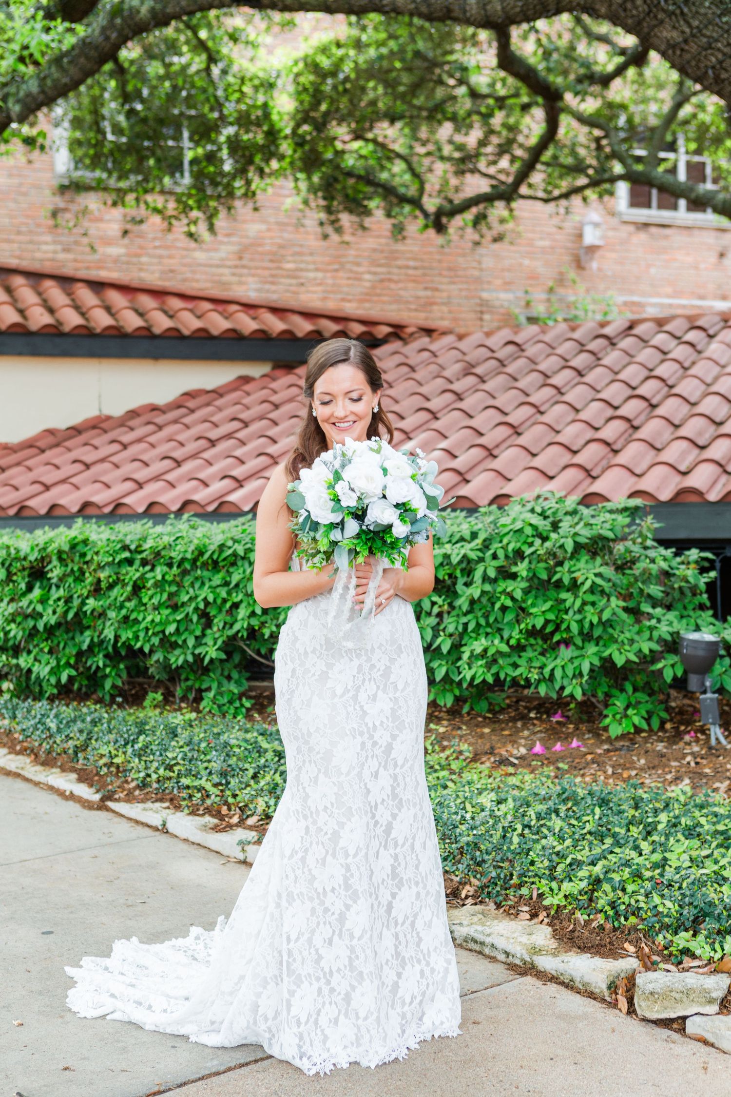 Bride in lace gown holding bouquet outdoors with tile roof and greenery.
