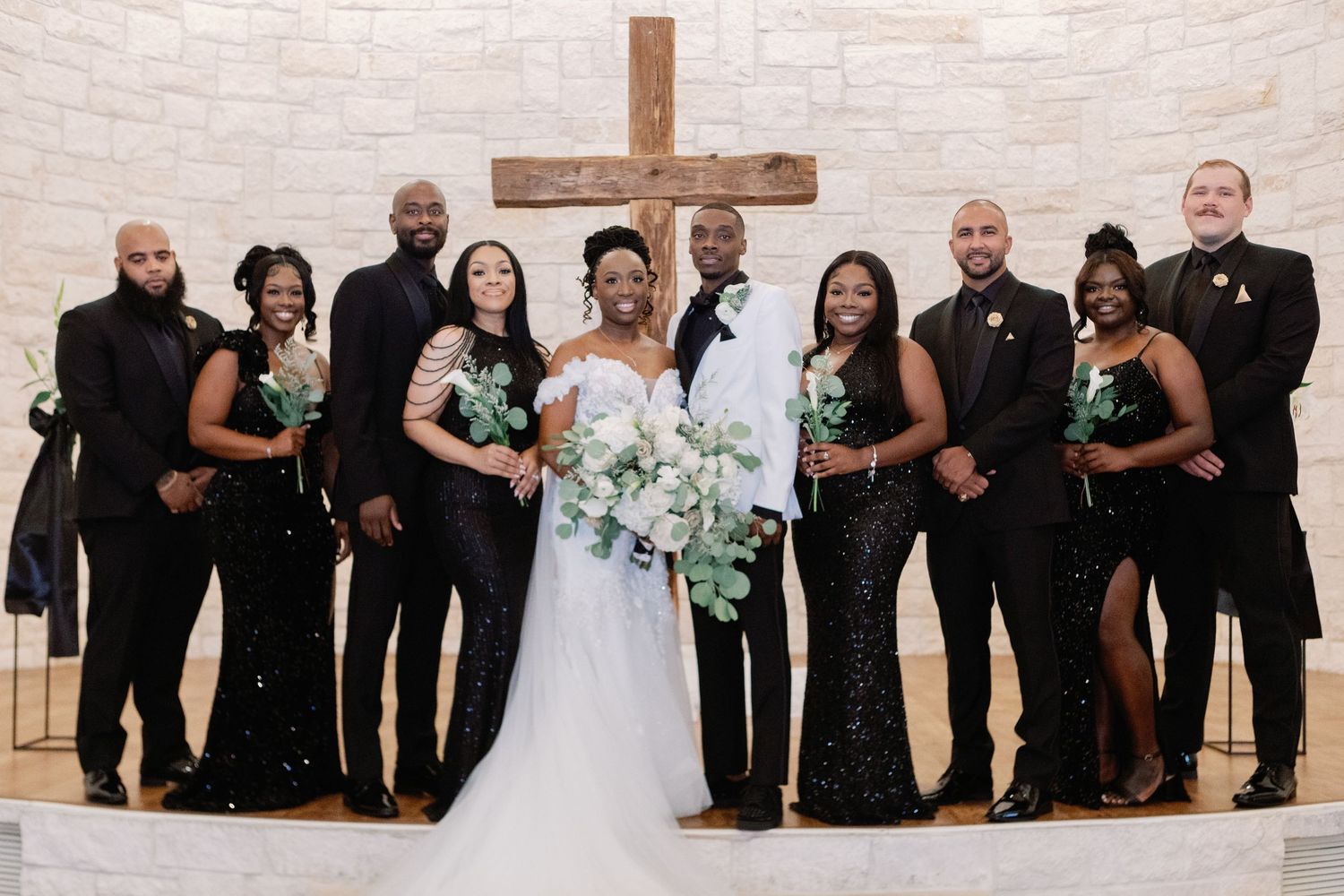 Wedding party in black attire poses against white brick wall with wooden cross during formal ceremony.
