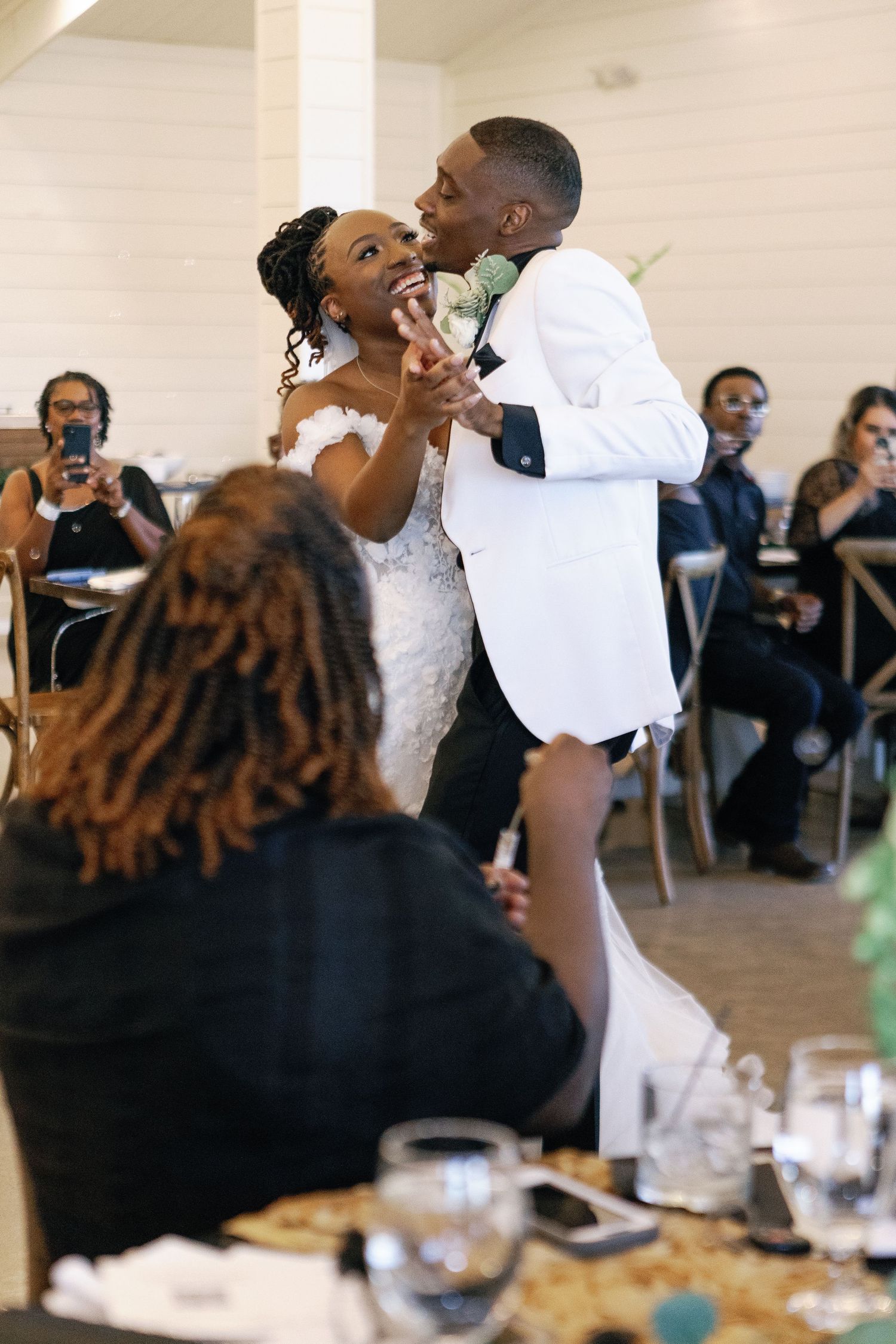 Couple shares a joyful moment during their first dance at wedding reception while guests look on and take photos.
