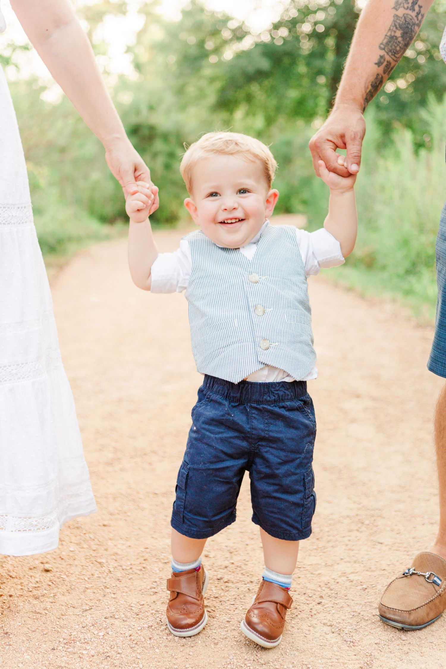 Toddler in blue vest and navy shorts holding hands while walking on dirt path.
