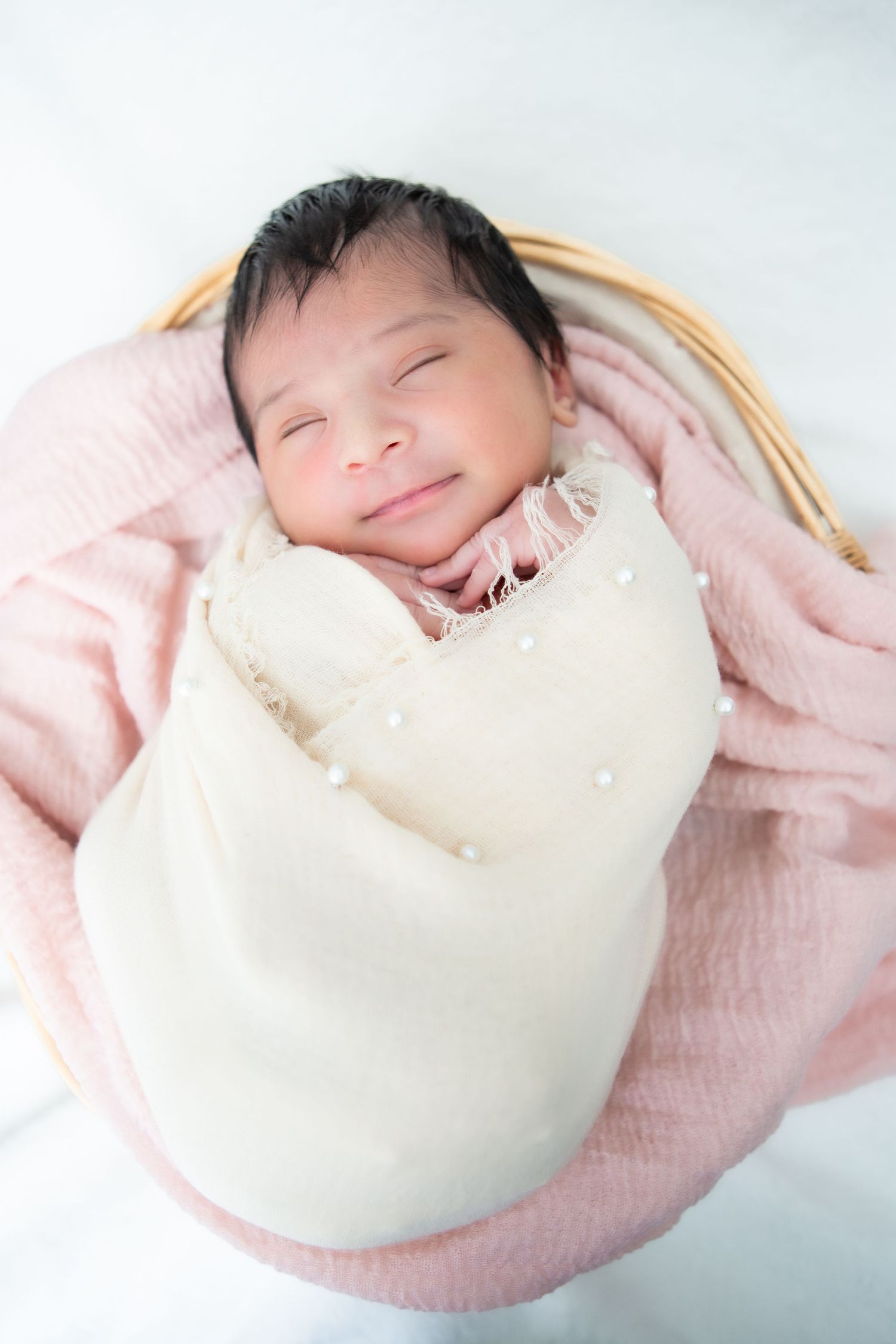 Infant in pink outfit nestled in cream-colored basket during studio photoshoot.