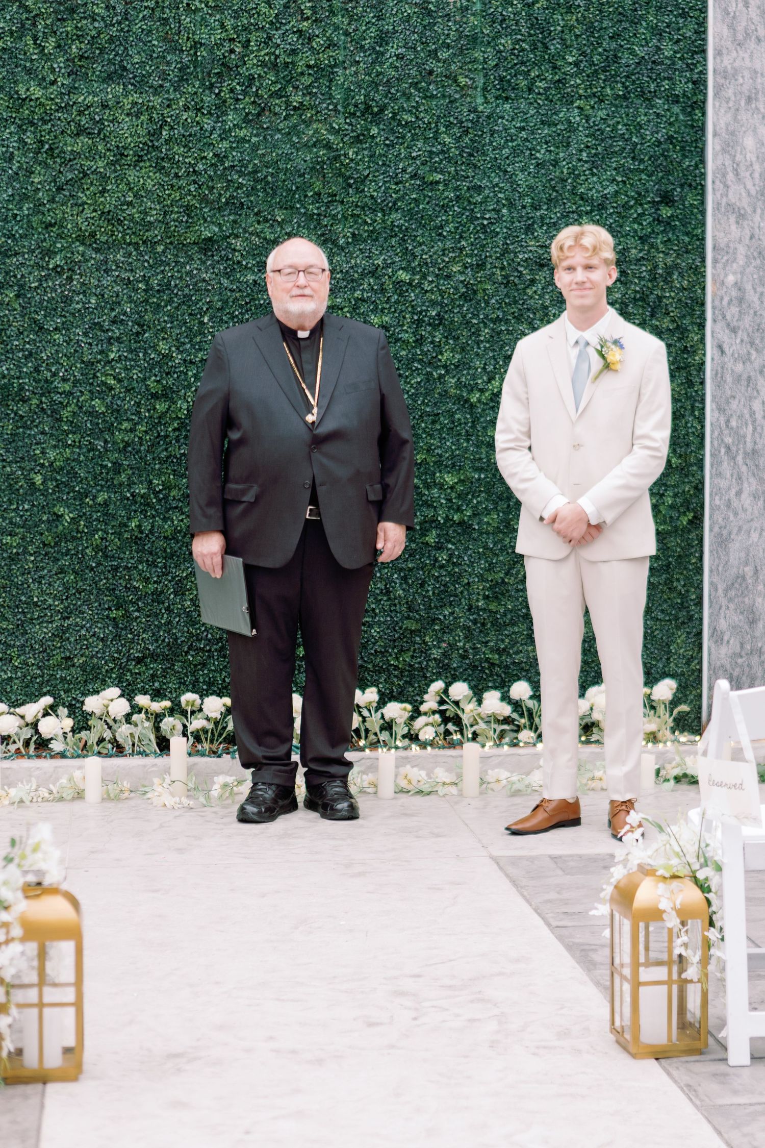 Two wedding attendants pose together against a lush green hedge wall with gold lanterns on the ground.