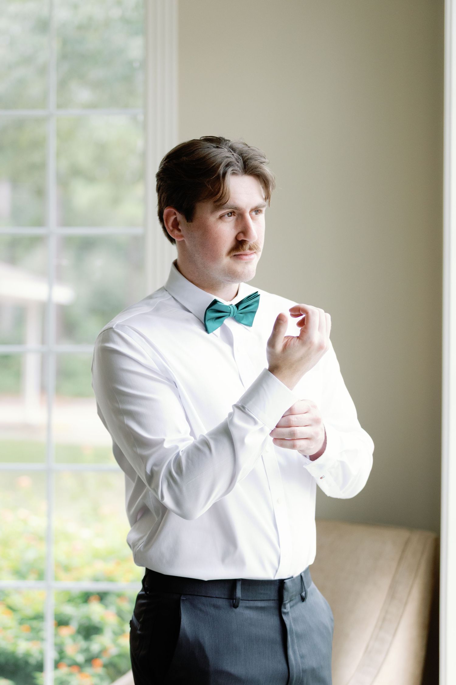 A groom in a white dress shirt and teal bow tie adjusts his cuffs while getting ready near a window.