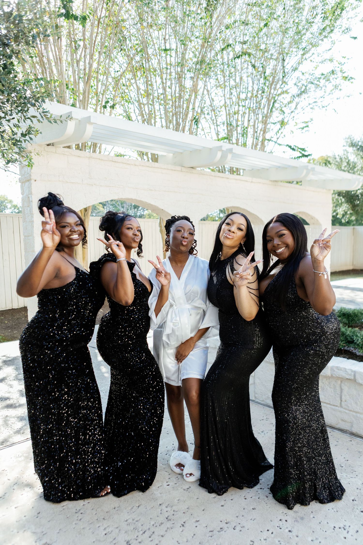 Bridal party in black dresses poses together outdoors with joyful expressions under a white tent.