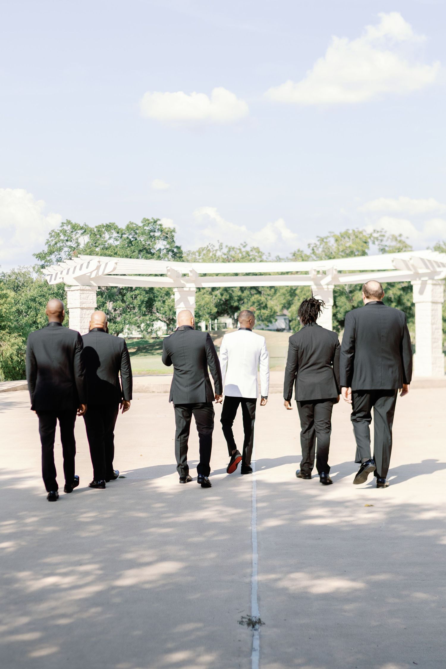 Groomsmen in black suits walking in a line on a sunny outdoor patio.