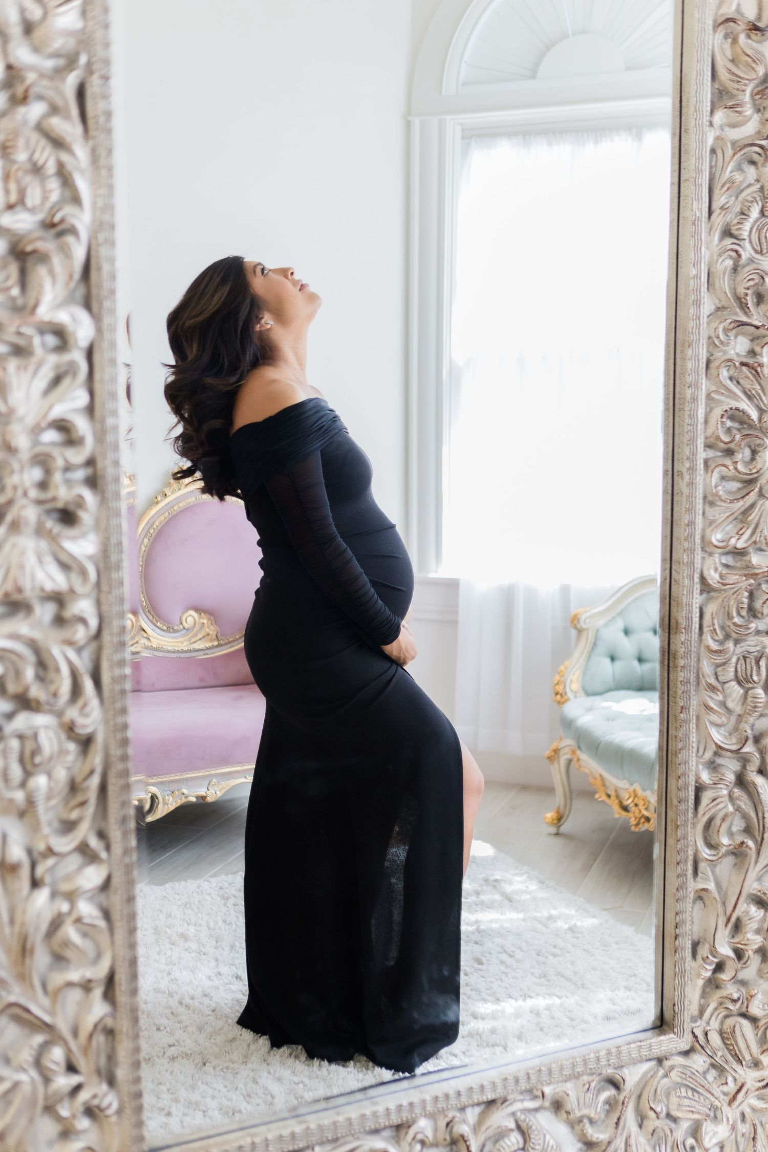 Side view of expecting mother in form-fitting black dress reflected in decorative silver mirror.