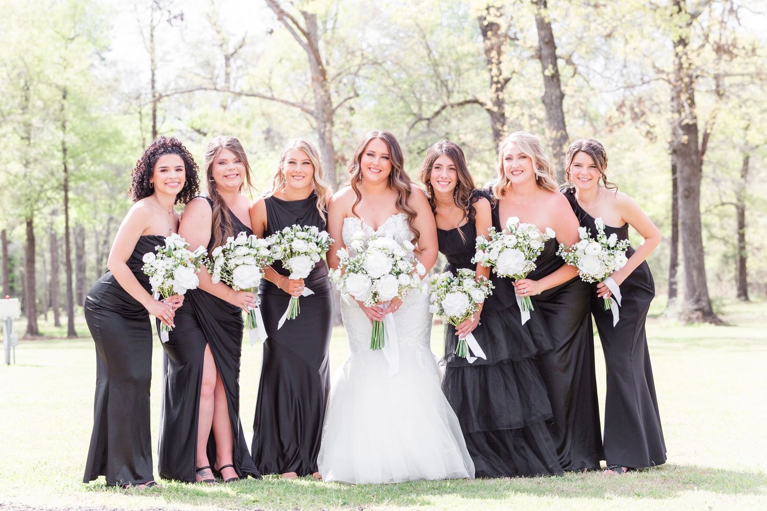 Wedding party in black dresses posing together outdoors among trees.