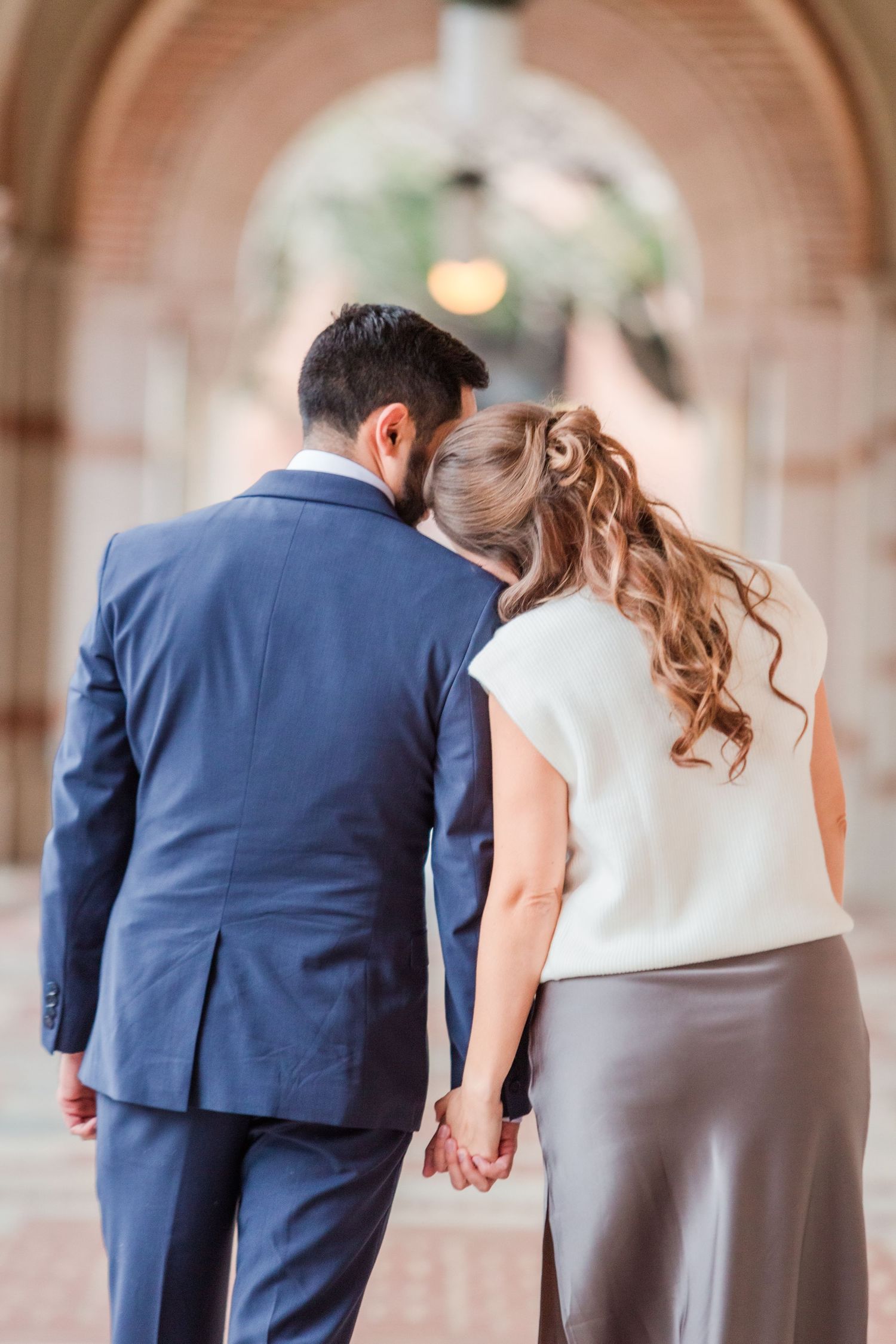 Couple holding hands strolls beneath cream-colored arches in hallway.