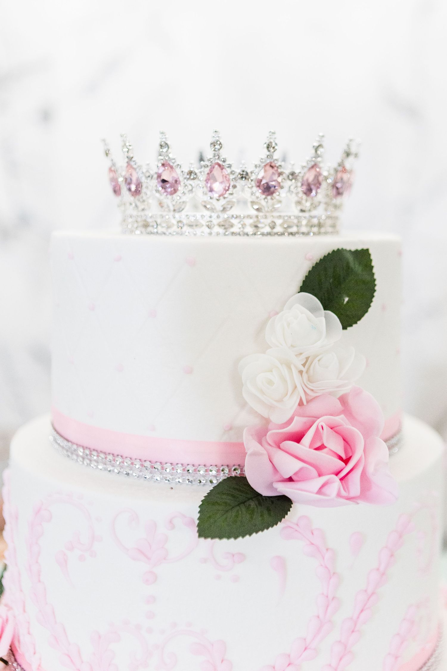 Elegant white tiered cake topped with crystal crown and pink rose decorations.