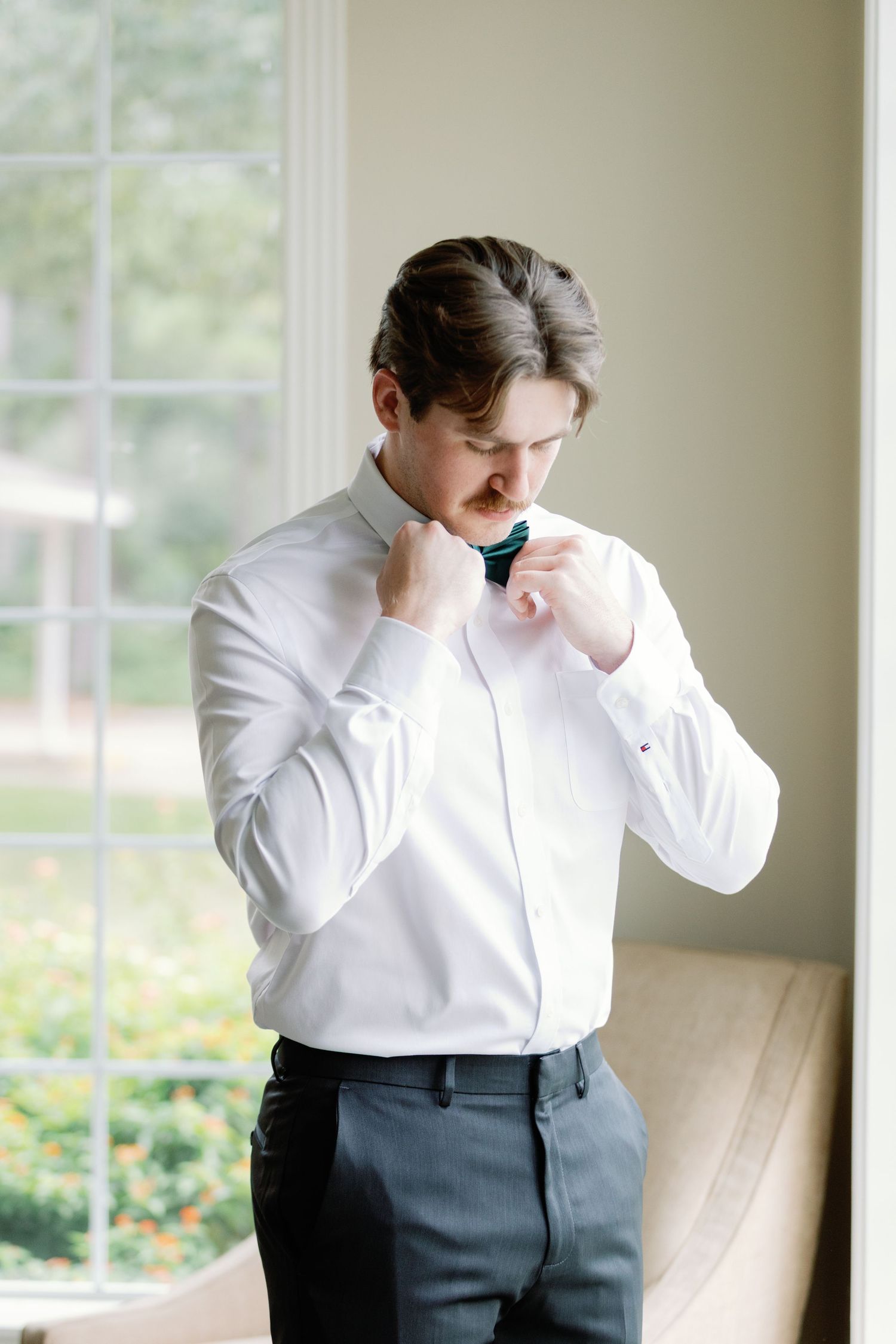 Groom in white shirt and dark pants adjusts his collar while standing near bright windows.