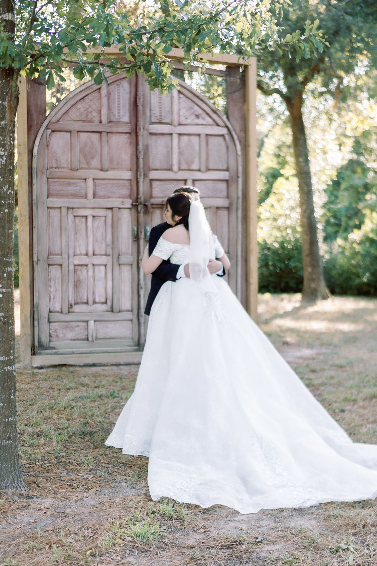 Bride in flowing white gown stands before rustic wooden doors in garden setting.