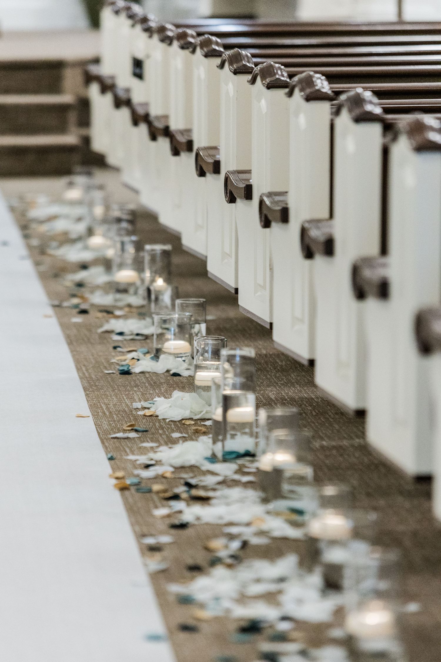 White church pews with scattered petals and candles line a wedding ceremony aisle.