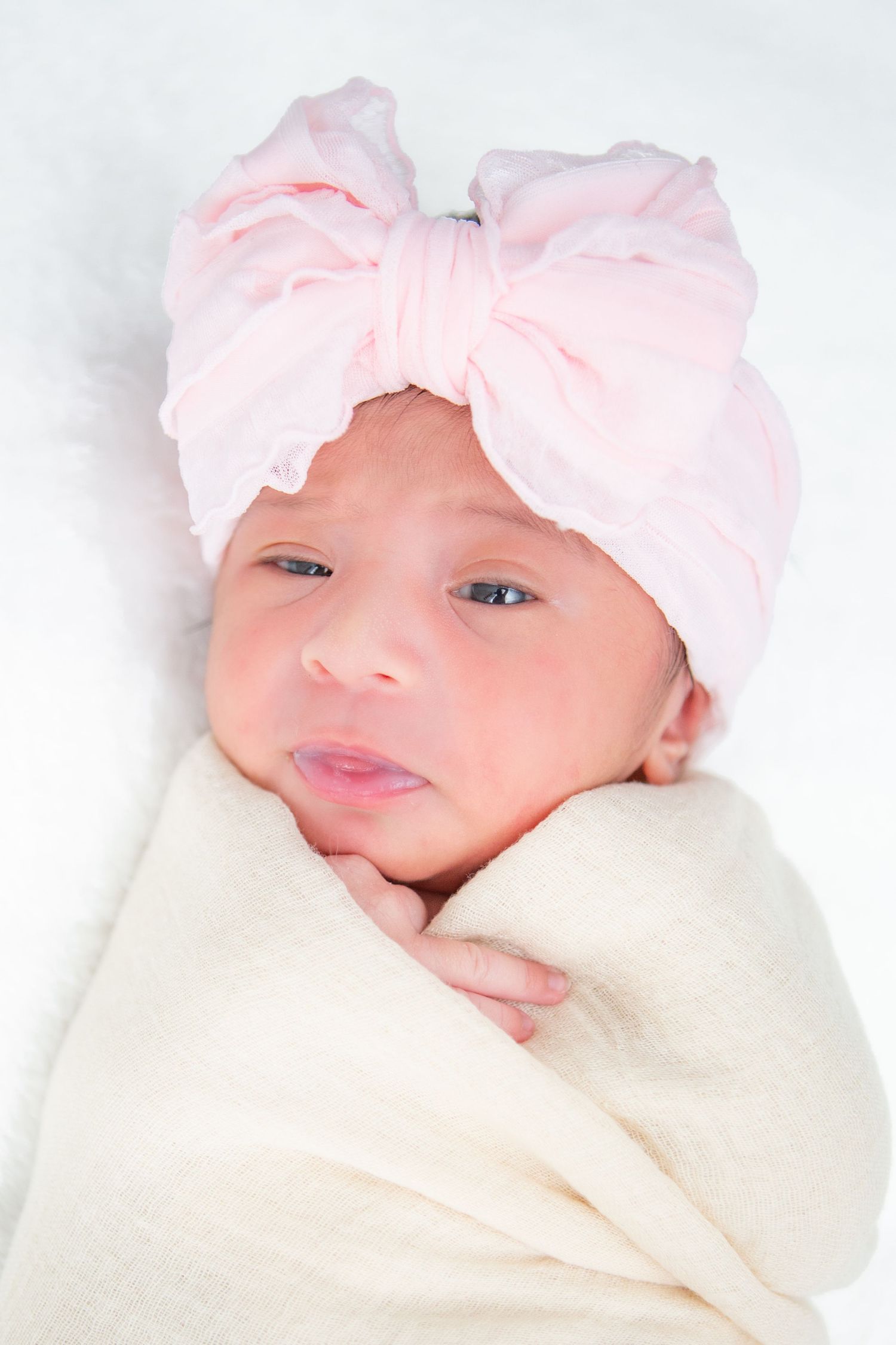 Baby wearing pink bonnet swaddled in soft cream blanket during professional photo session.