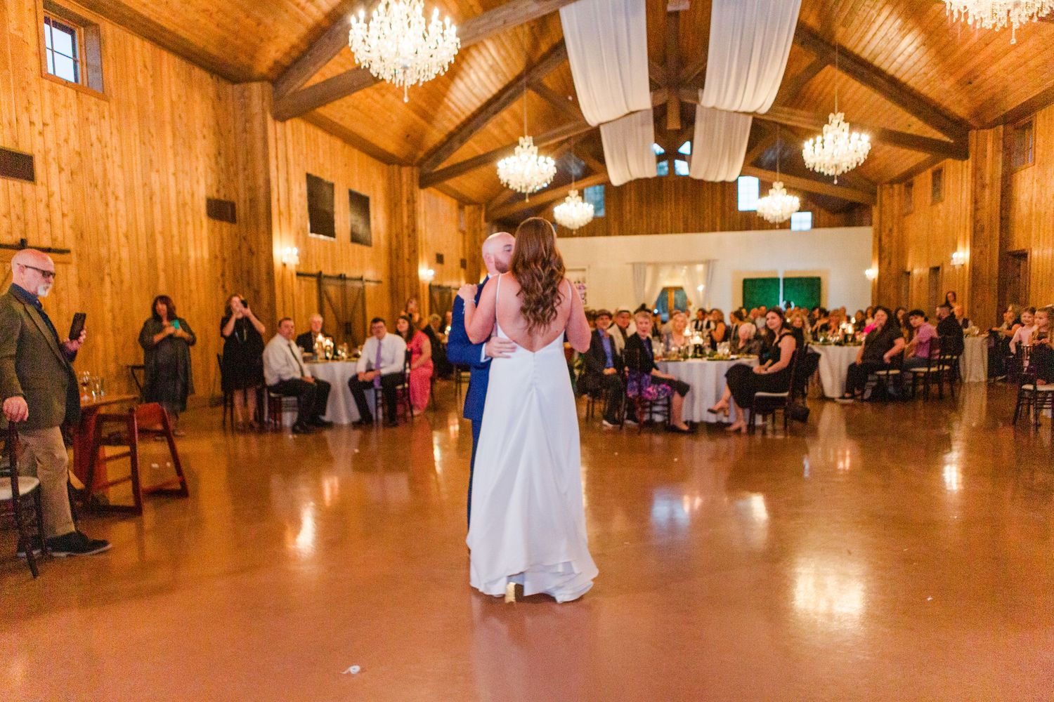 Panoramic view of a rustic barn wedding reception with wooden walls and crystal chandeliers.