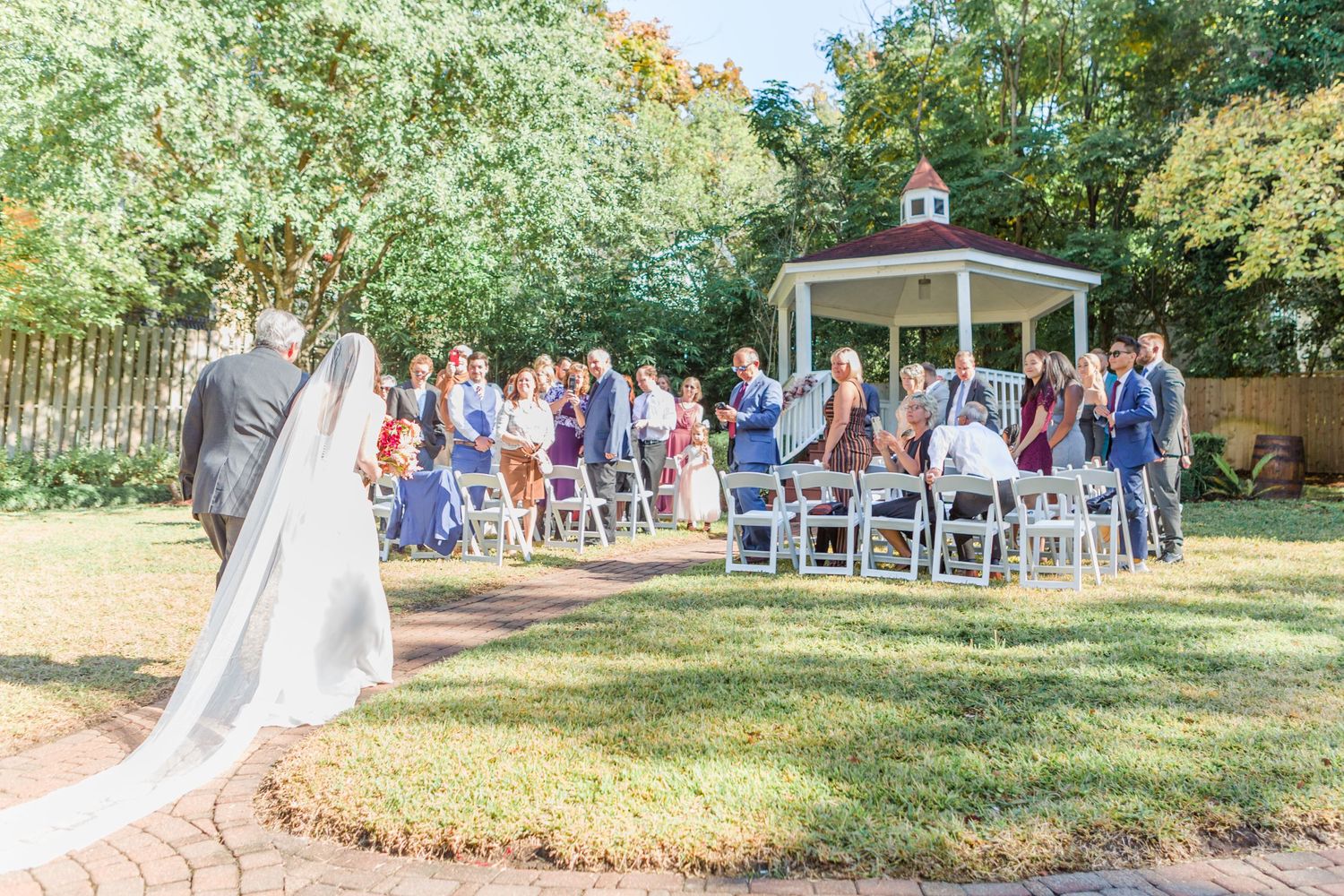 Wedding ceremony site with white chairs arranged on lawn facing gazebo under trees.