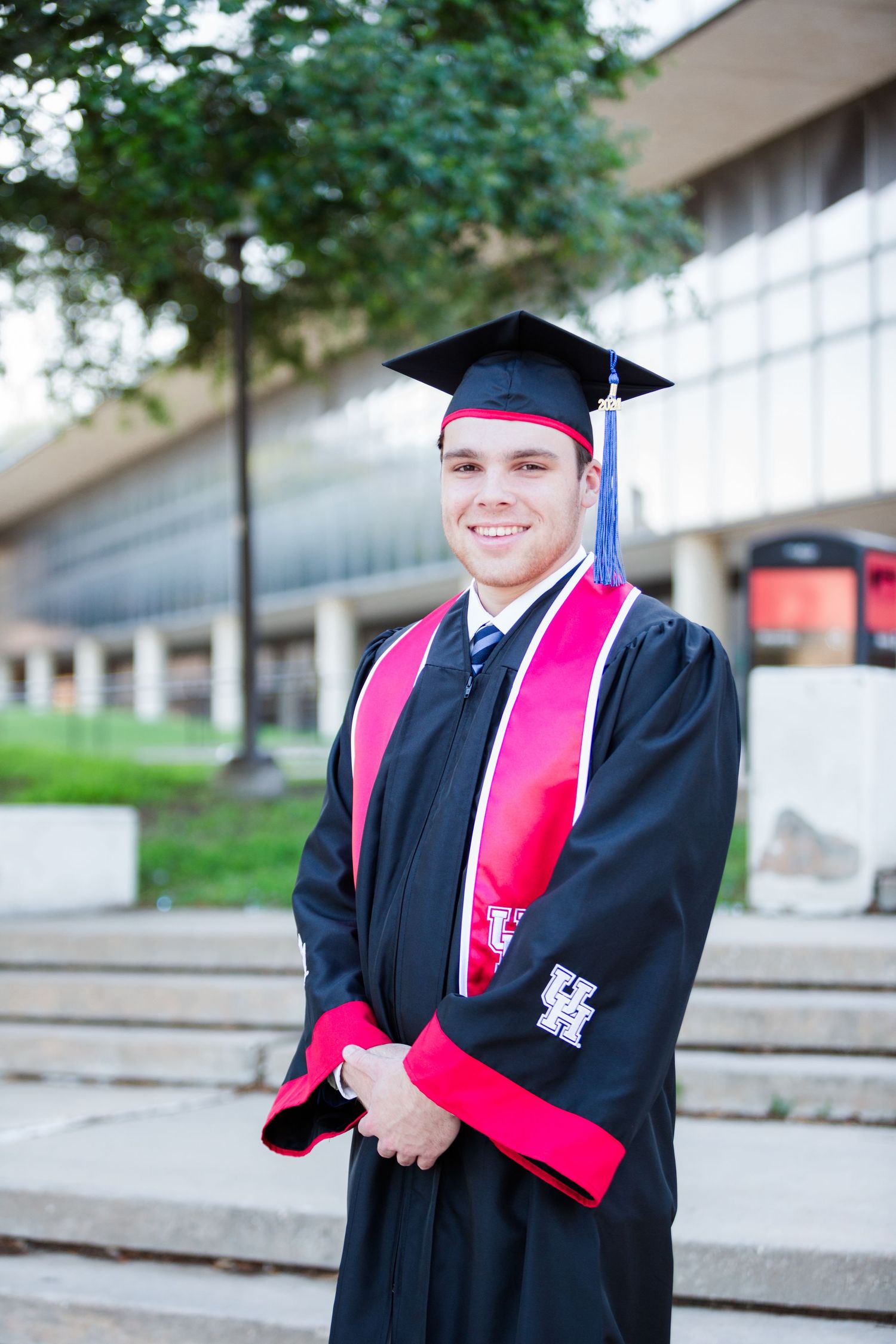Graduate in black cap and gown with pink stole poses on concrete steps outdoors.