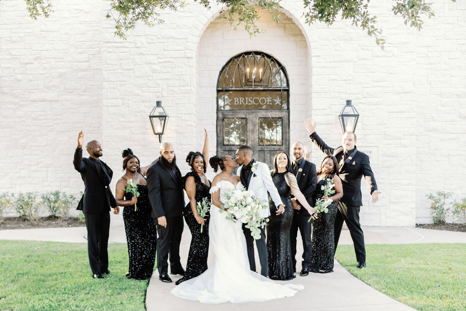 Wedding party poses for playful group photos outside stone church with raised arms celebrating joyfully.