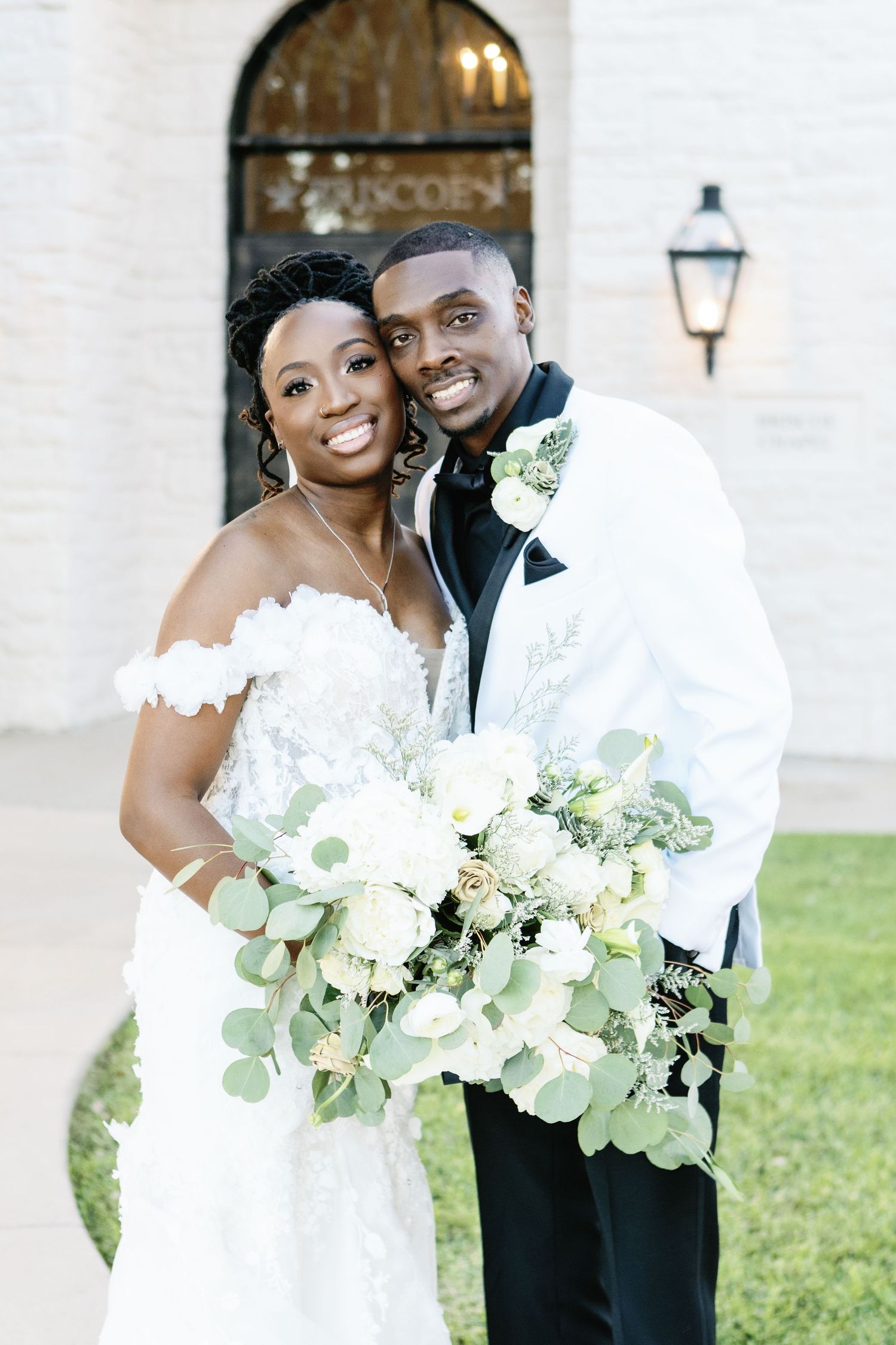 A joyful wedding couple embraces while holding an elegant white floral bouquet with greenery.