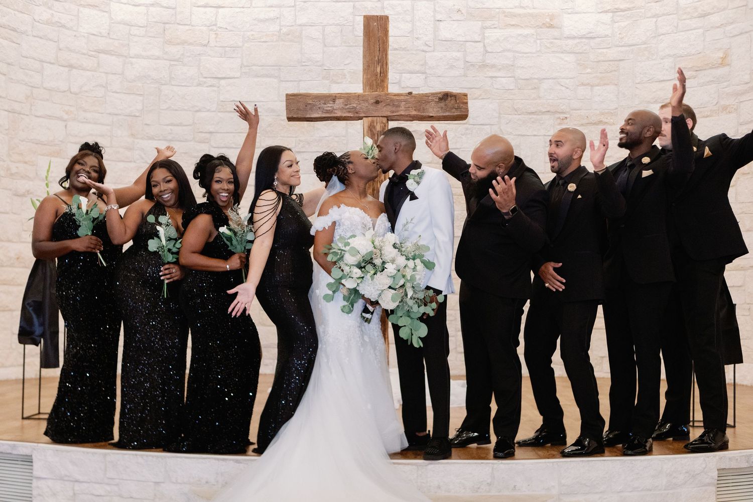 Wedding celebration with bridal party posing against white brick wall with wooden cross.