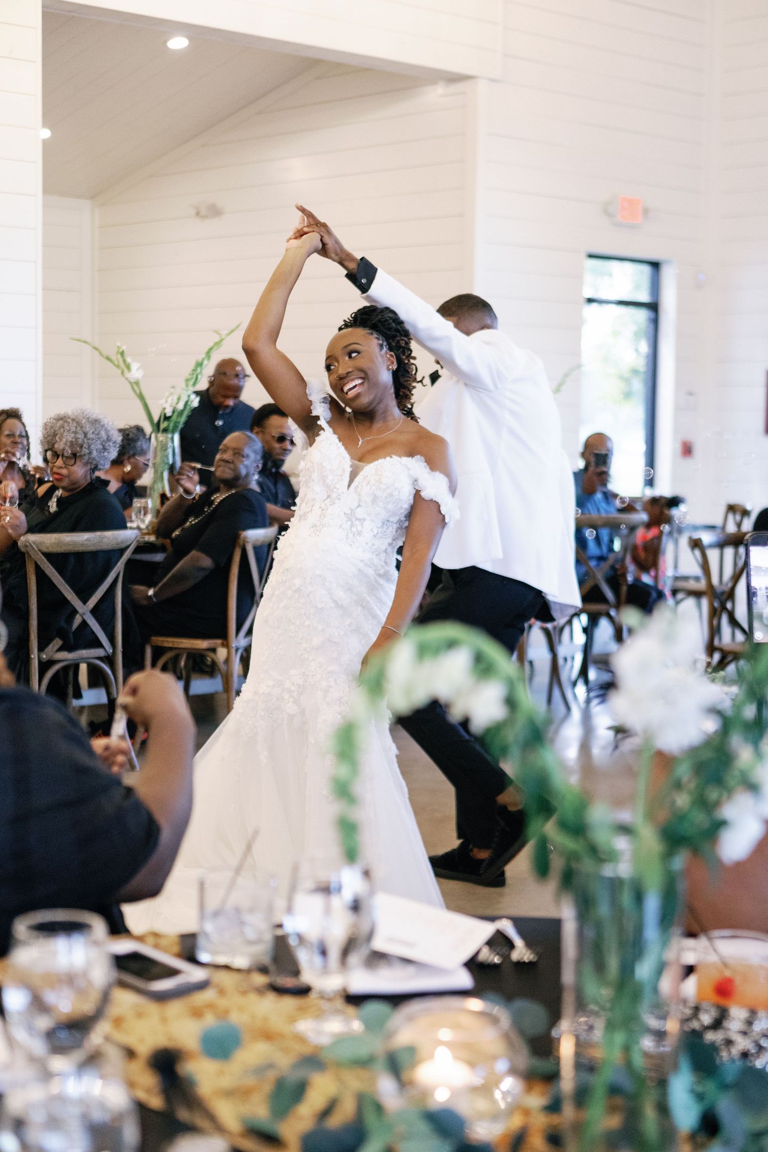 Newlyweds share a celebratory dance during their wedding reception in an elegant white room decorated with greenery.